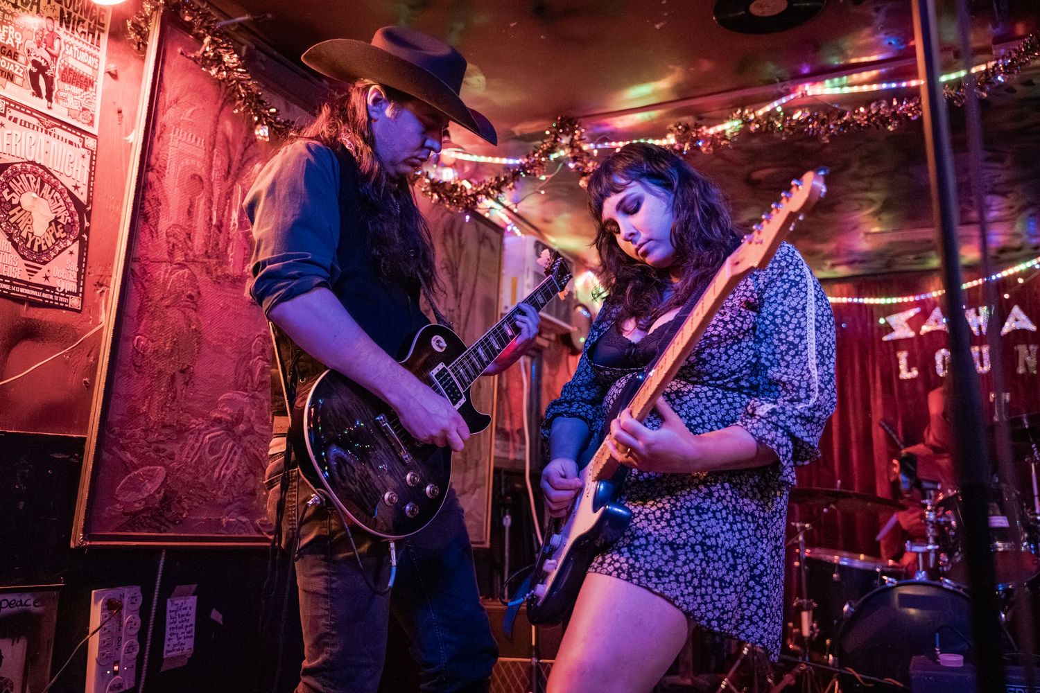 Musicians perform on stage in a dimly lit venue bathed in vibrant purple and red lighting during a live show.