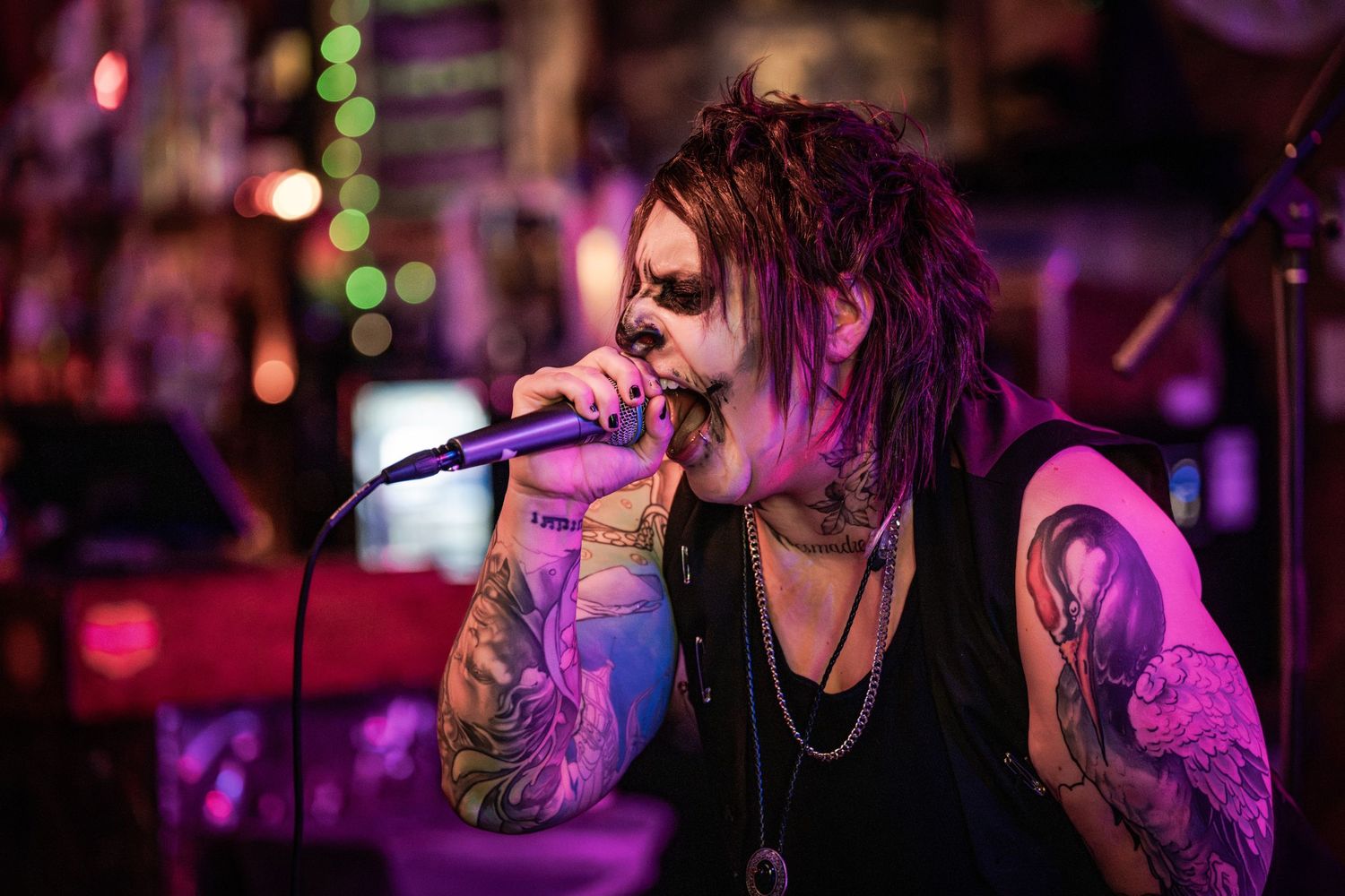 A performer with long hair sings passionately into a microphone on stage under dramatic purple lighting.