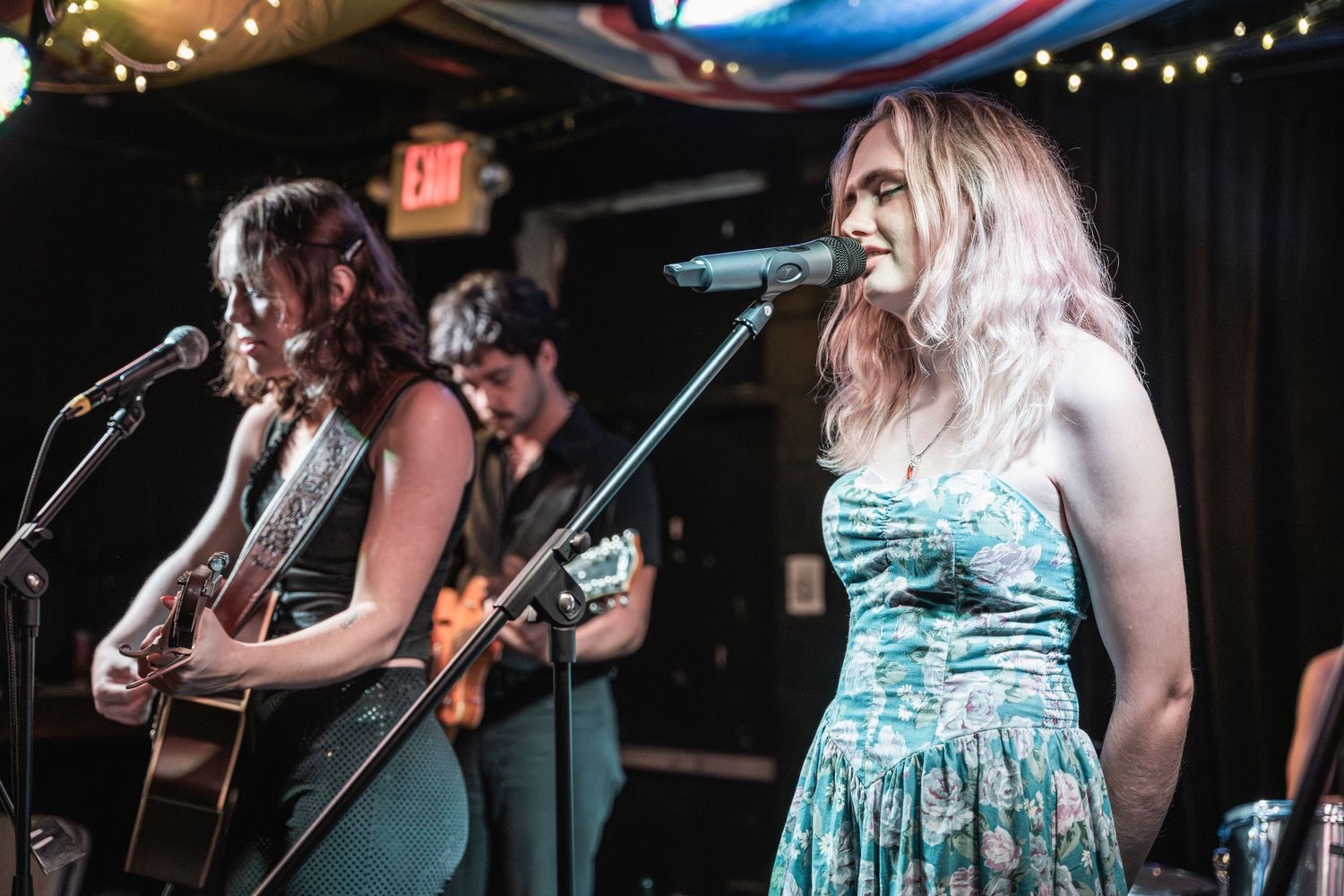 Musicians perform on stage with acoustic guitars and string instruments under twinkling lights at a live music venue.