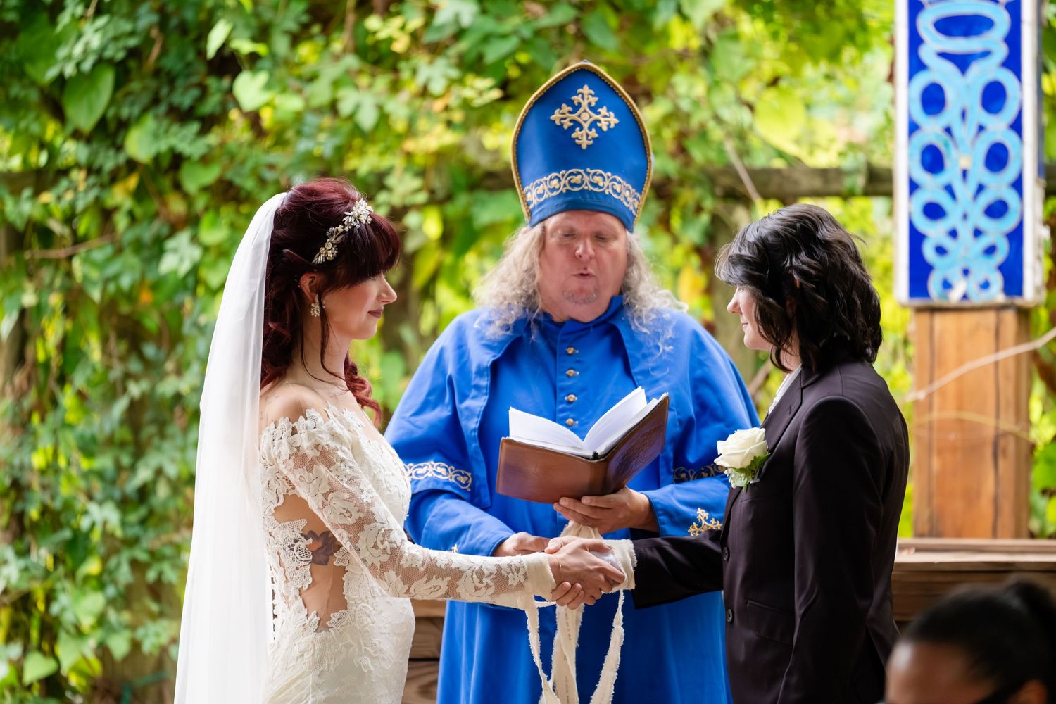 Outdoor wedding ceremony with officiant in blue shirt reading from book while couple stands together in garden setting.