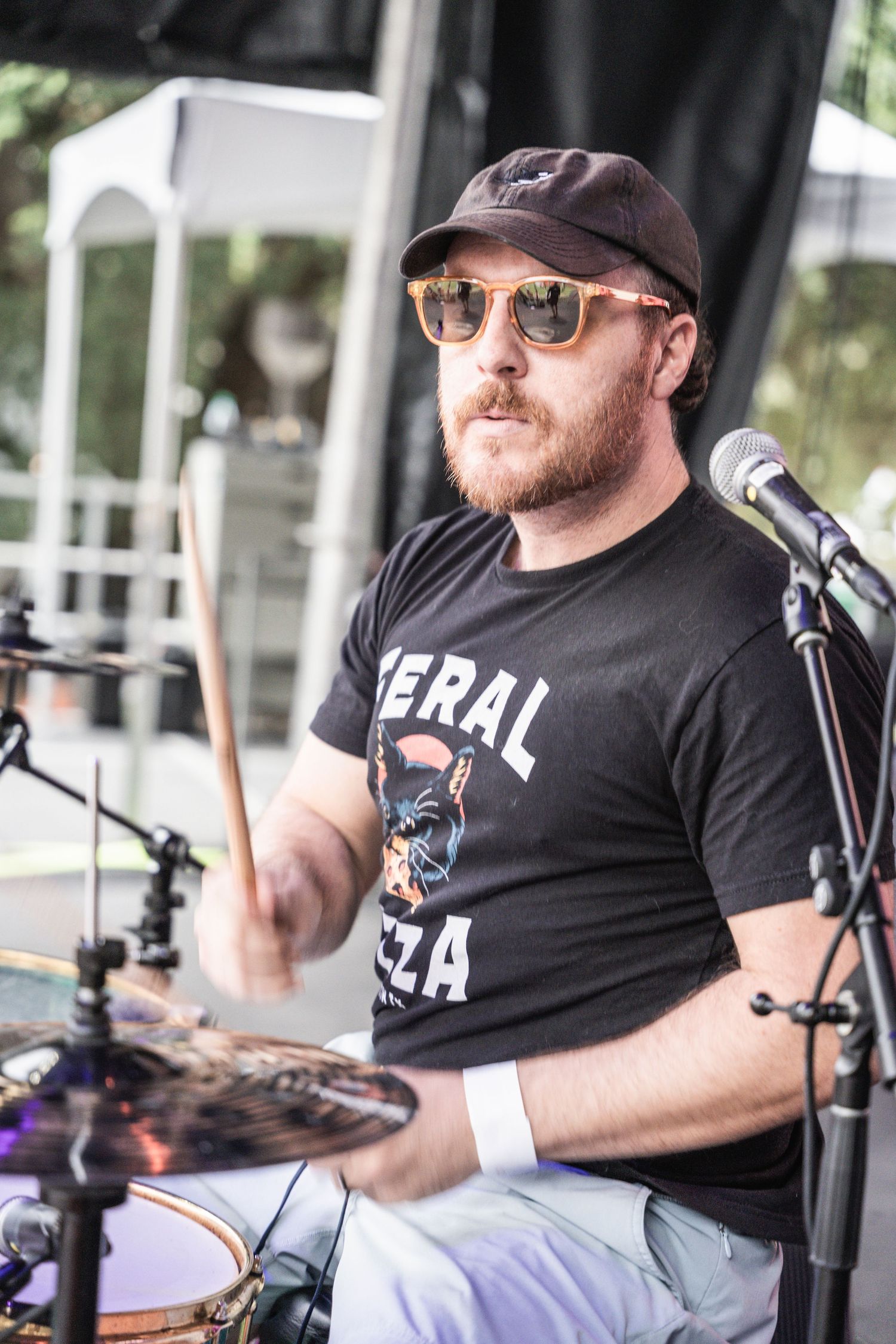 A drummer wearing sunglasses and a black t-shirt performs behind a drum kit at an outdoor venue.