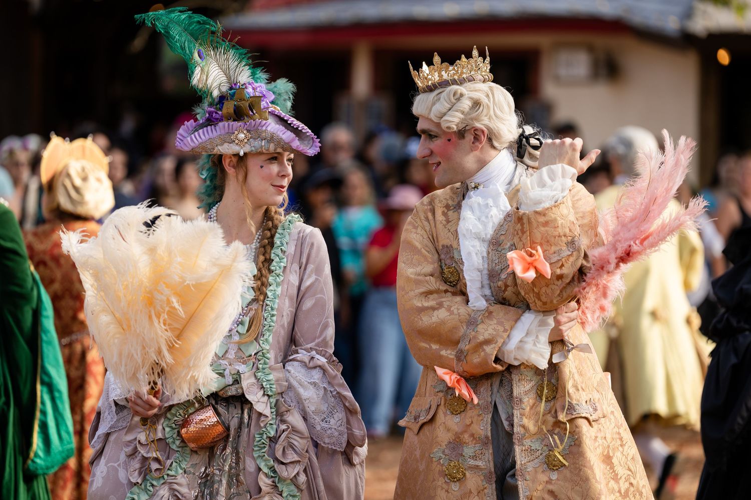 People in ornate 18th century period costumes with powdered wigs and elaborate dresses socialize at a historical reenactment event.
