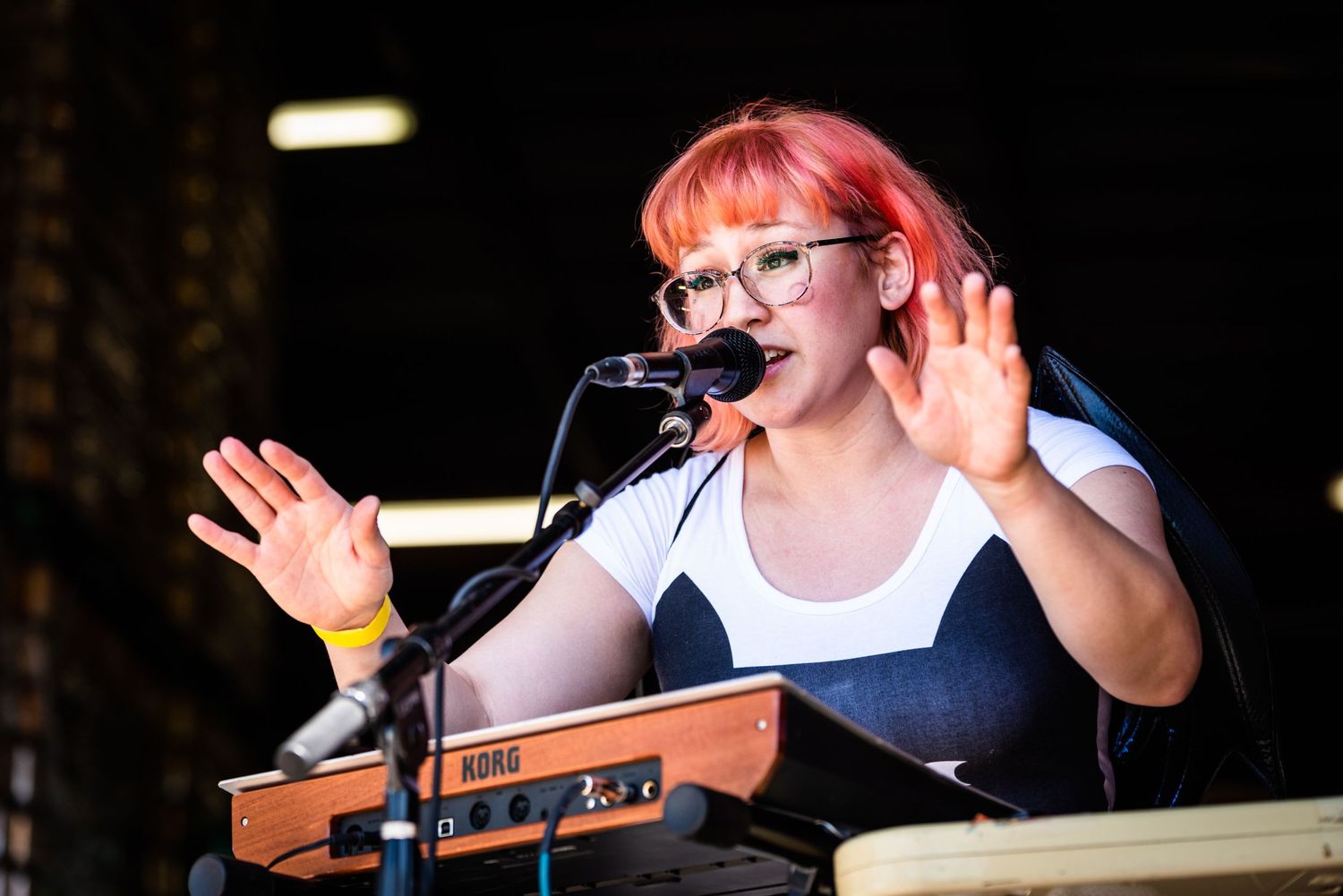 A person with pink hair and glasses performs on stage with expressive hand gestures.