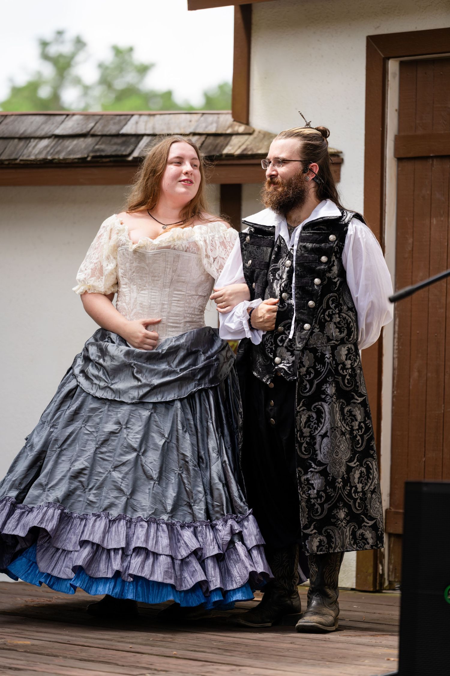Two performers in Renaissance faire costumes, a woman in gray gown and man in ornate vest, walk together on outdoor stage.