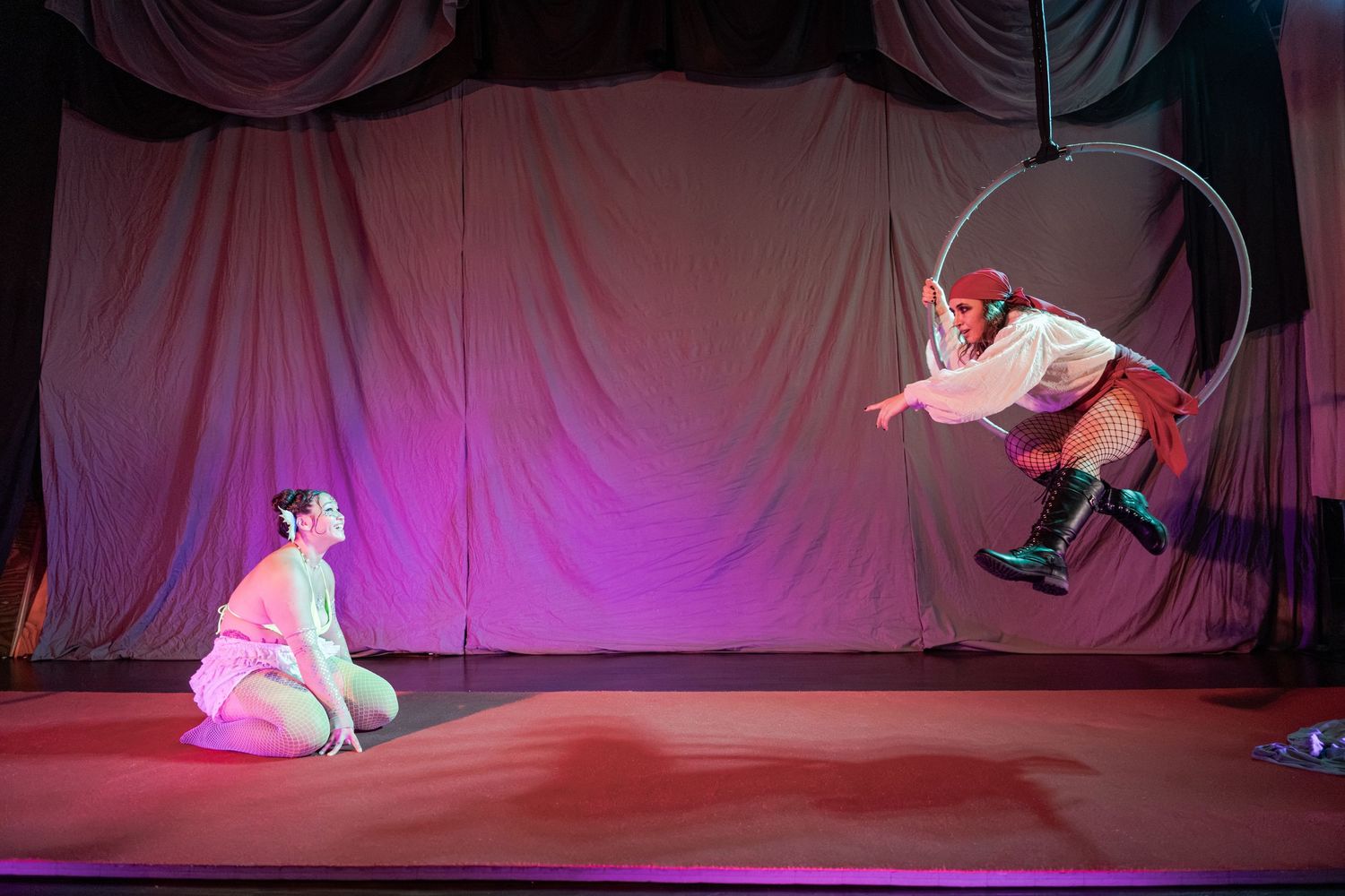 Aerial acrobat performs on a hoop above a stage while another performer kneels below in a theatrical circus show.