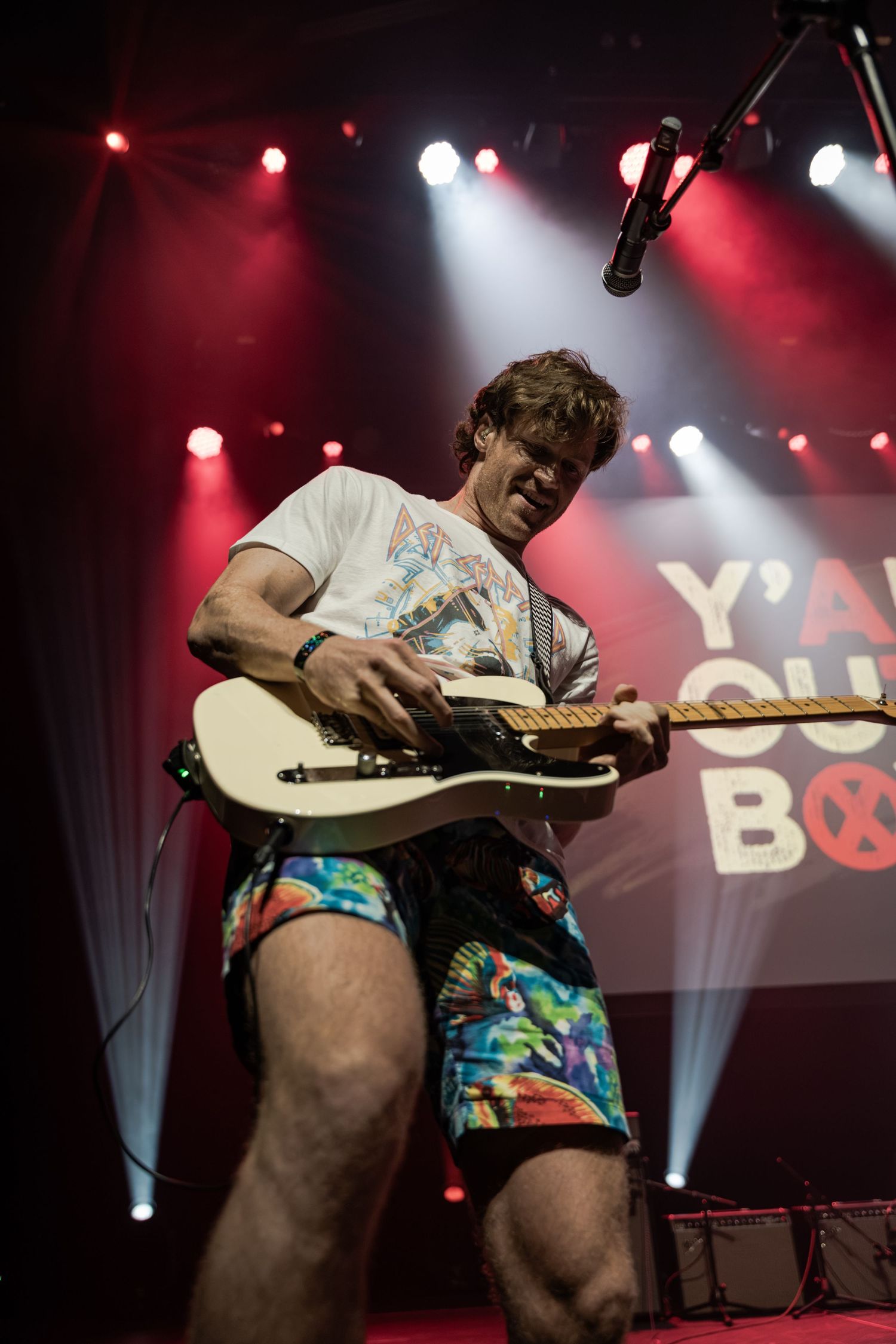 Guitarist performing on stage in colorful shorts and white tee, playing electric guitar under red and white stage lights.