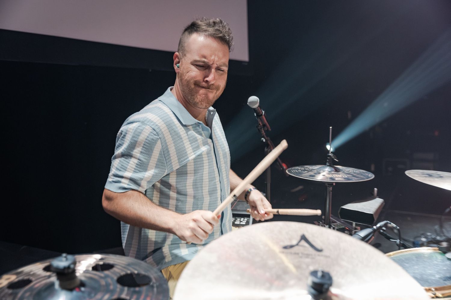 Smiling male drummer playing cymbals with drumsticks on stage under blue stage lighting.