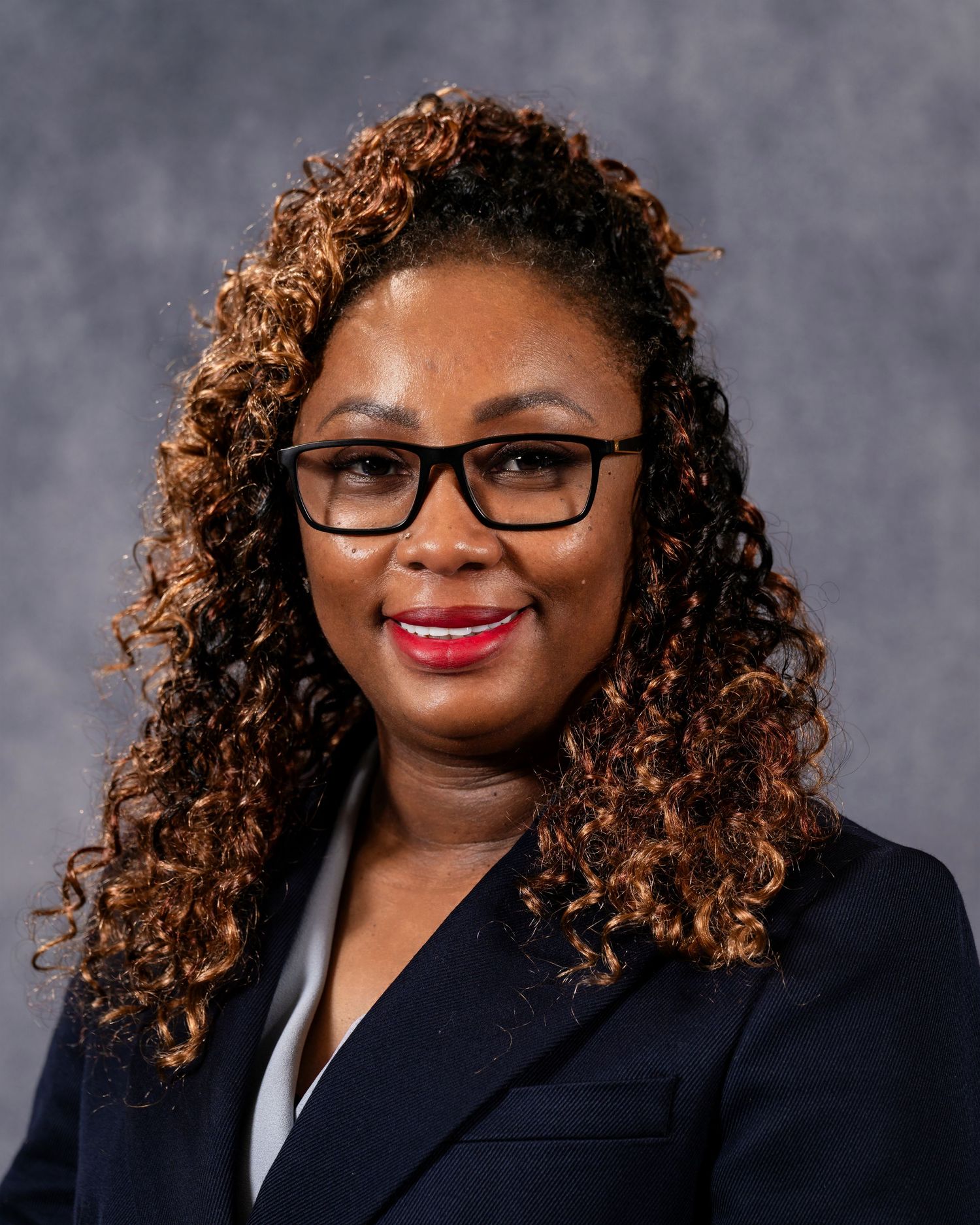 Professional headshot of a woman with curly auburn hair wearing black glasses and a navy blazer.