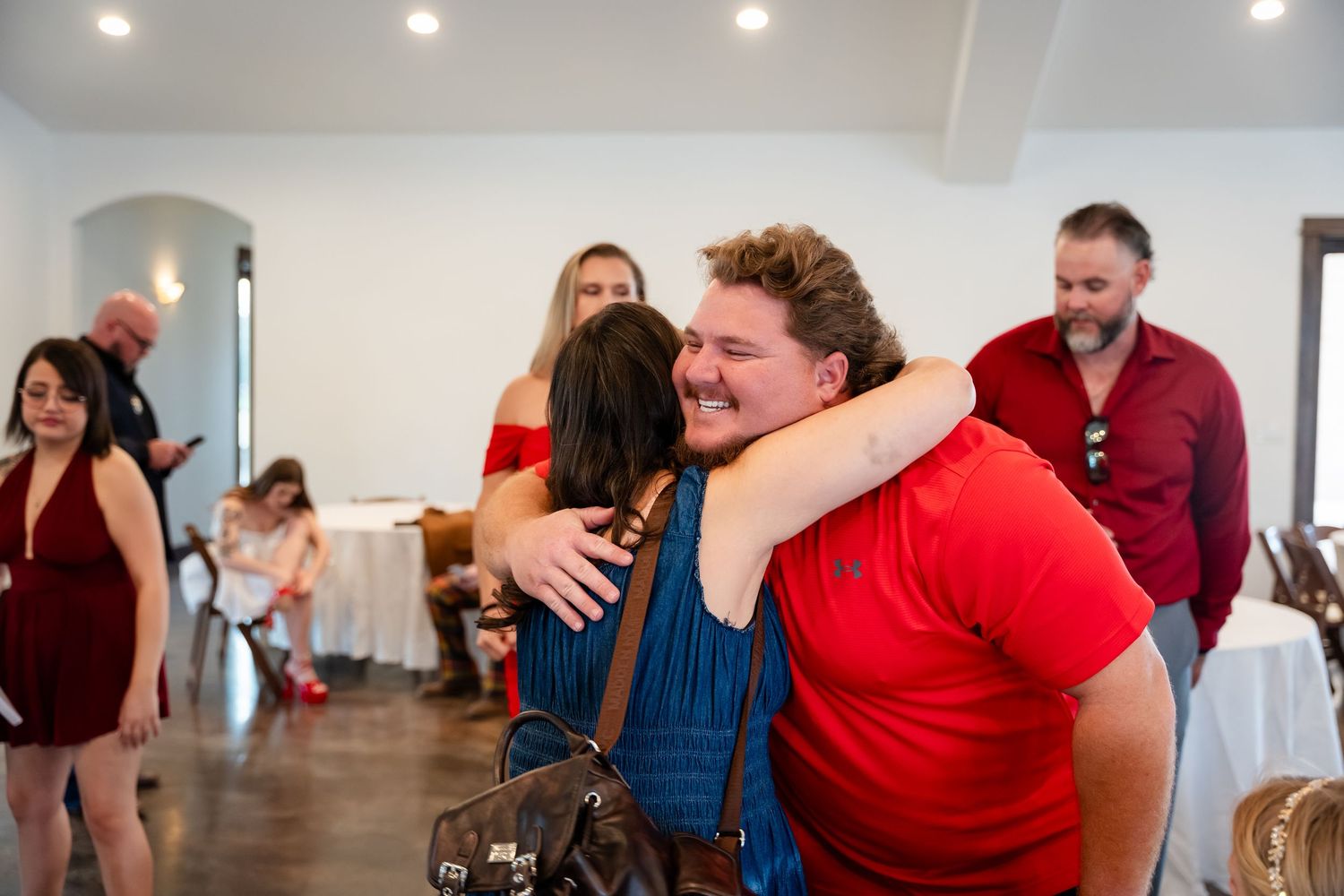 Two people sharing a warm hug at an indoor social gathering, with guests mingling in a bright, elegant venue.