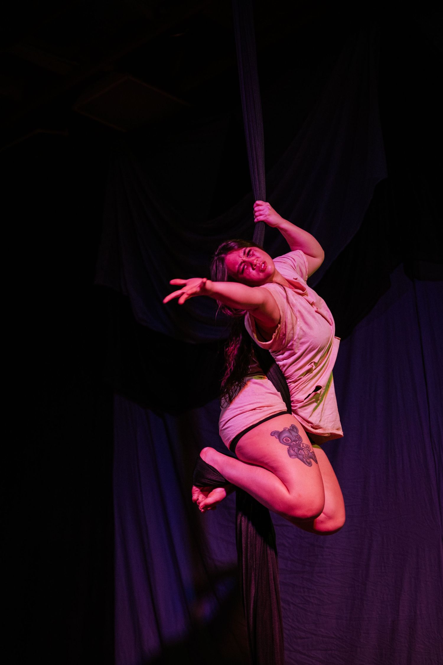 Aerial silk performer suspended mid-air under pink stage lighting during a dramatic circus arts performance.