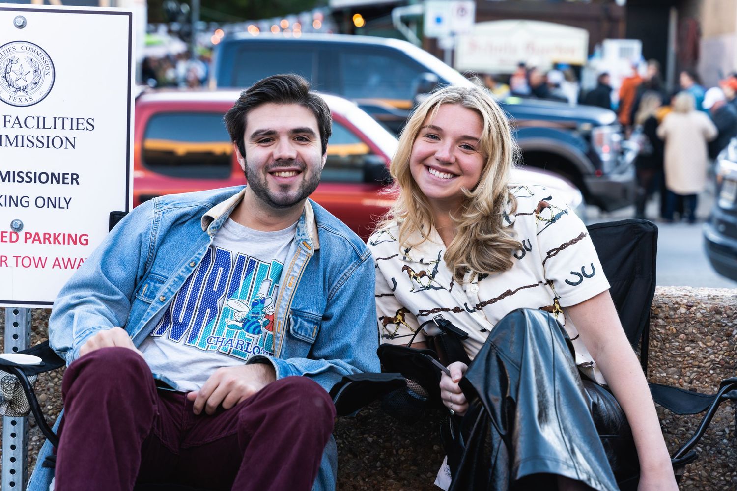 Two people sitting casually on a bench near event signage at dusk.
