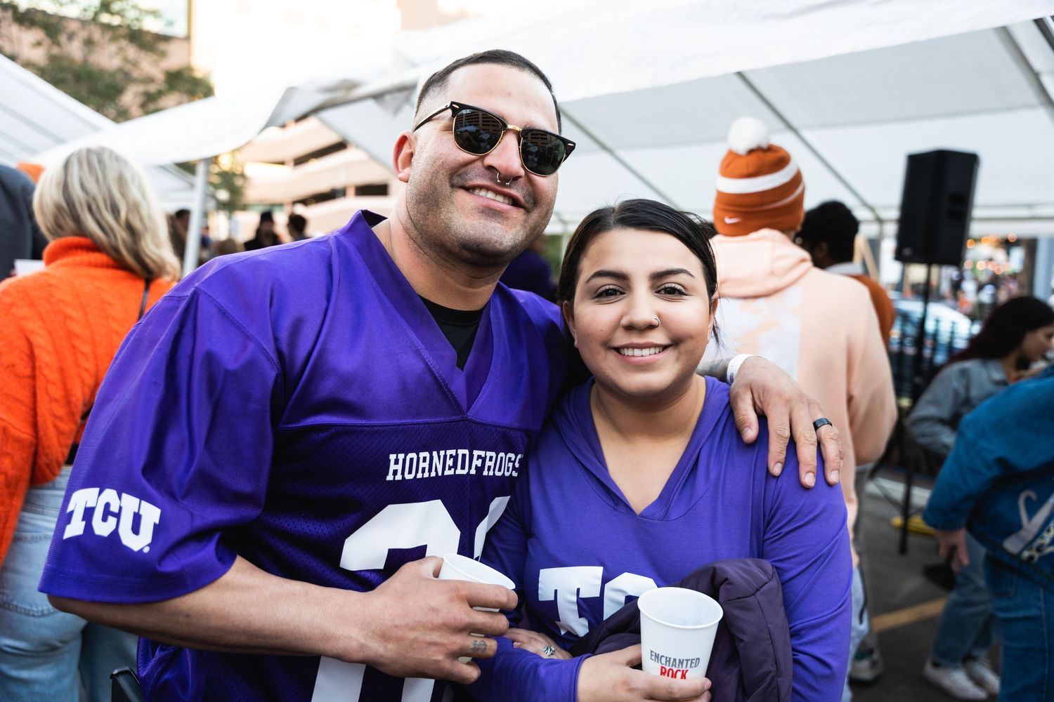 Fans wearing purple TCU gear smile while enjoying drinks at an outdoor event.