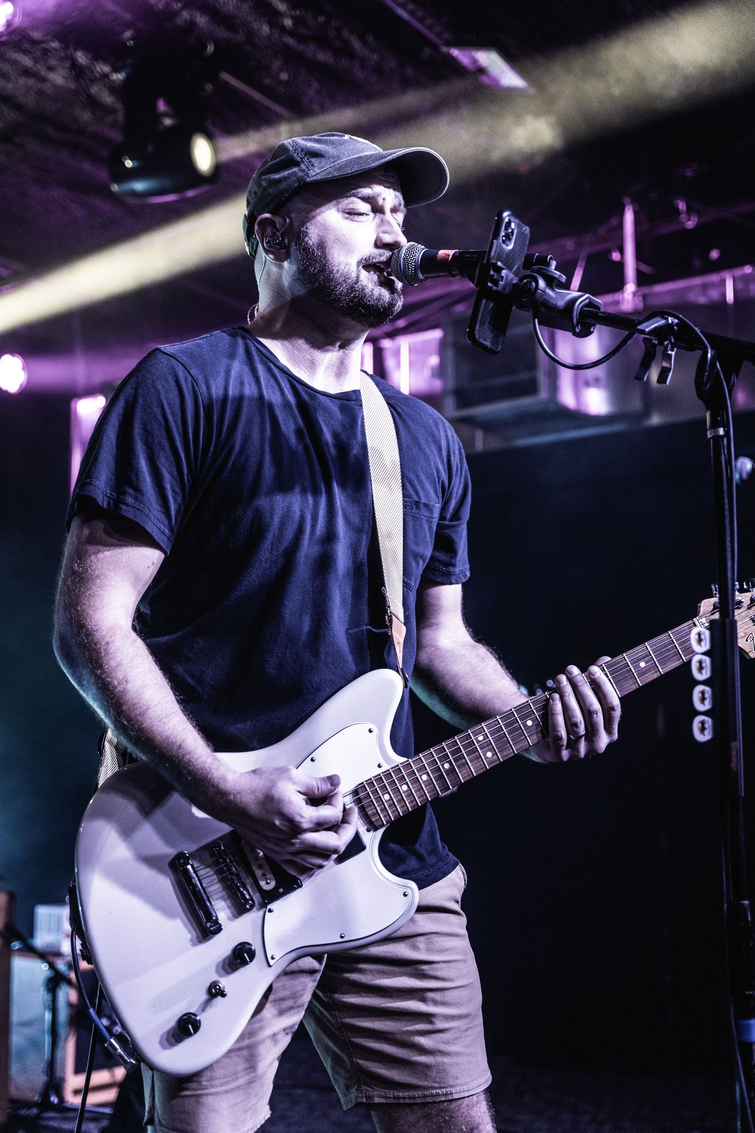 Musician in navy t-shirt performs with white electric guitar under dramatic purple stage lighting.