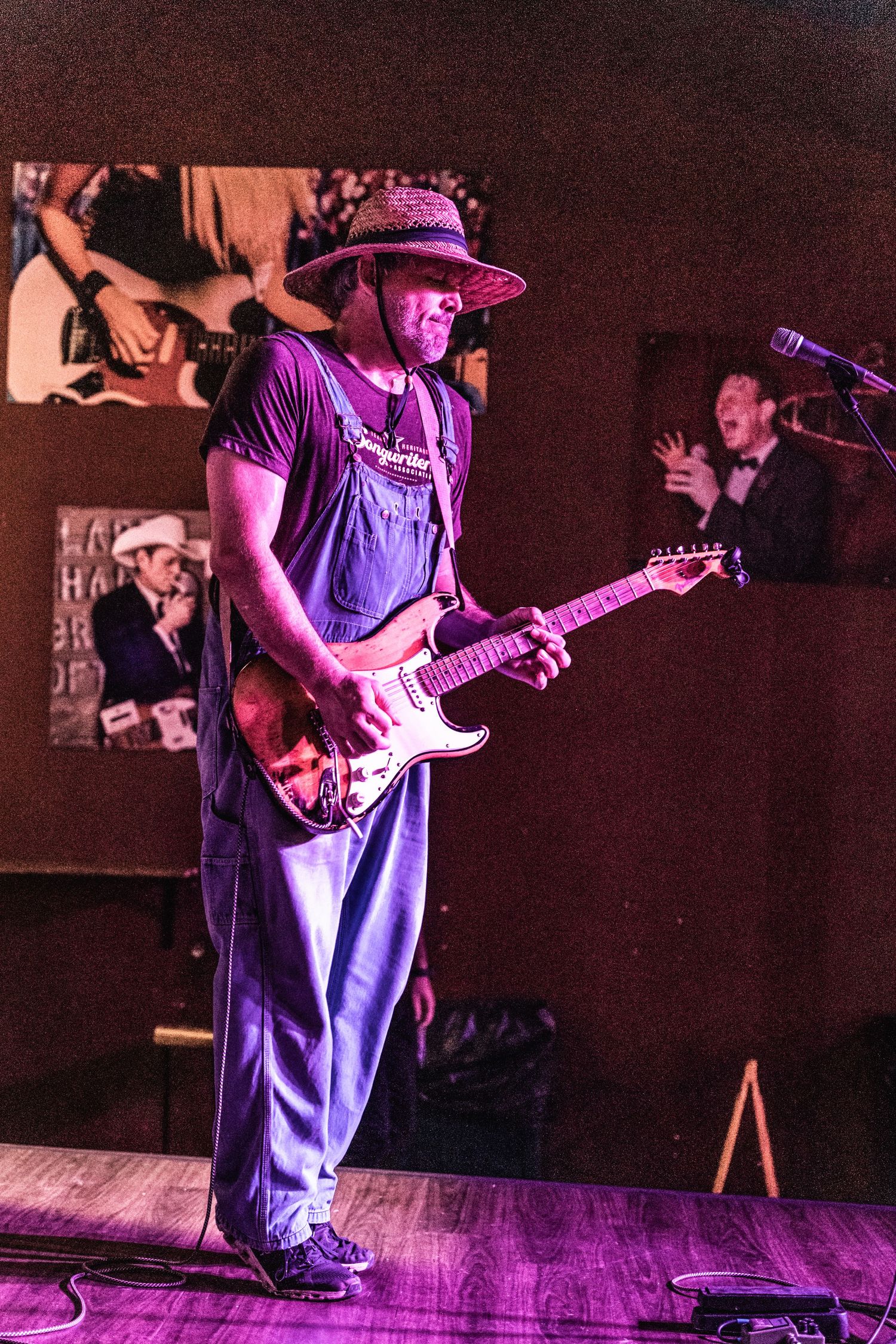 Guitar player wearing overalls and hat performs on stage with purple lighting effects.