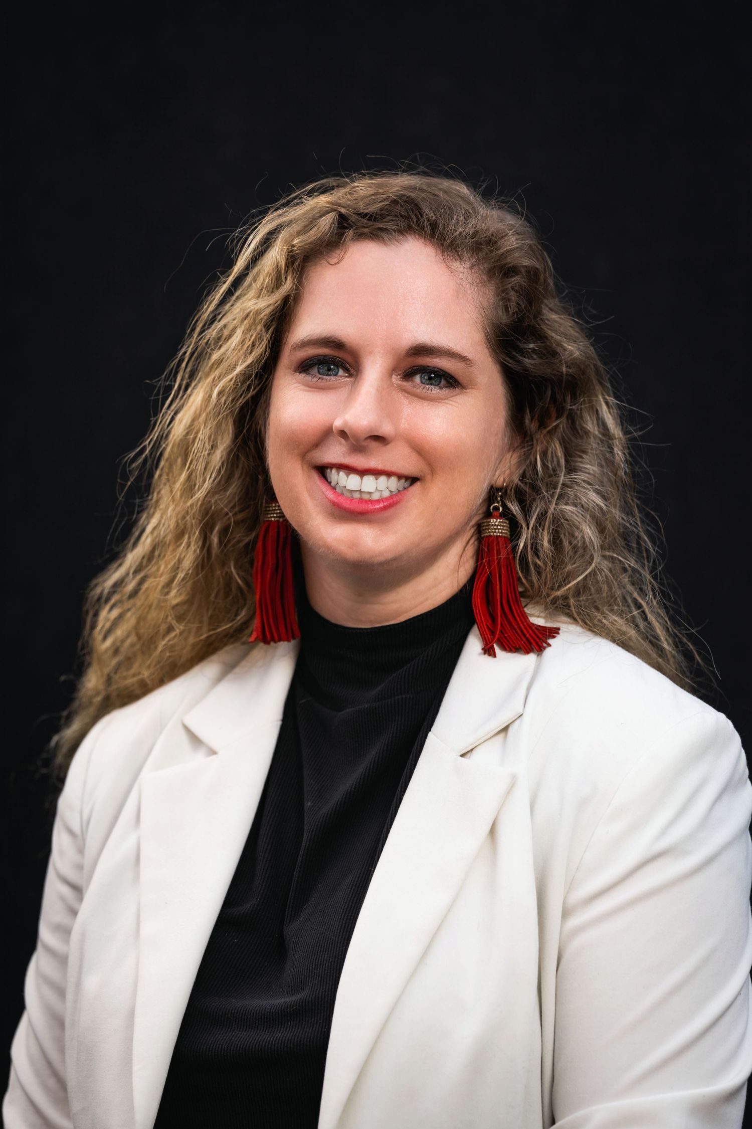 Portrait of woman in white blazer and red tassel earrings smiling against black background.