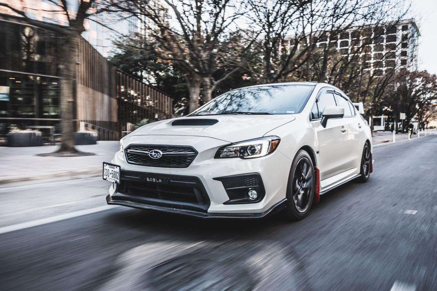 Lowered white Subaru WRX STI with custom wheels photographed in motion on city street.