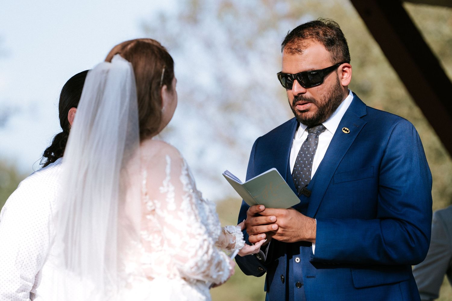 An officiant in sunglasses and blue suit conducts an outdoor wedding ceremony.