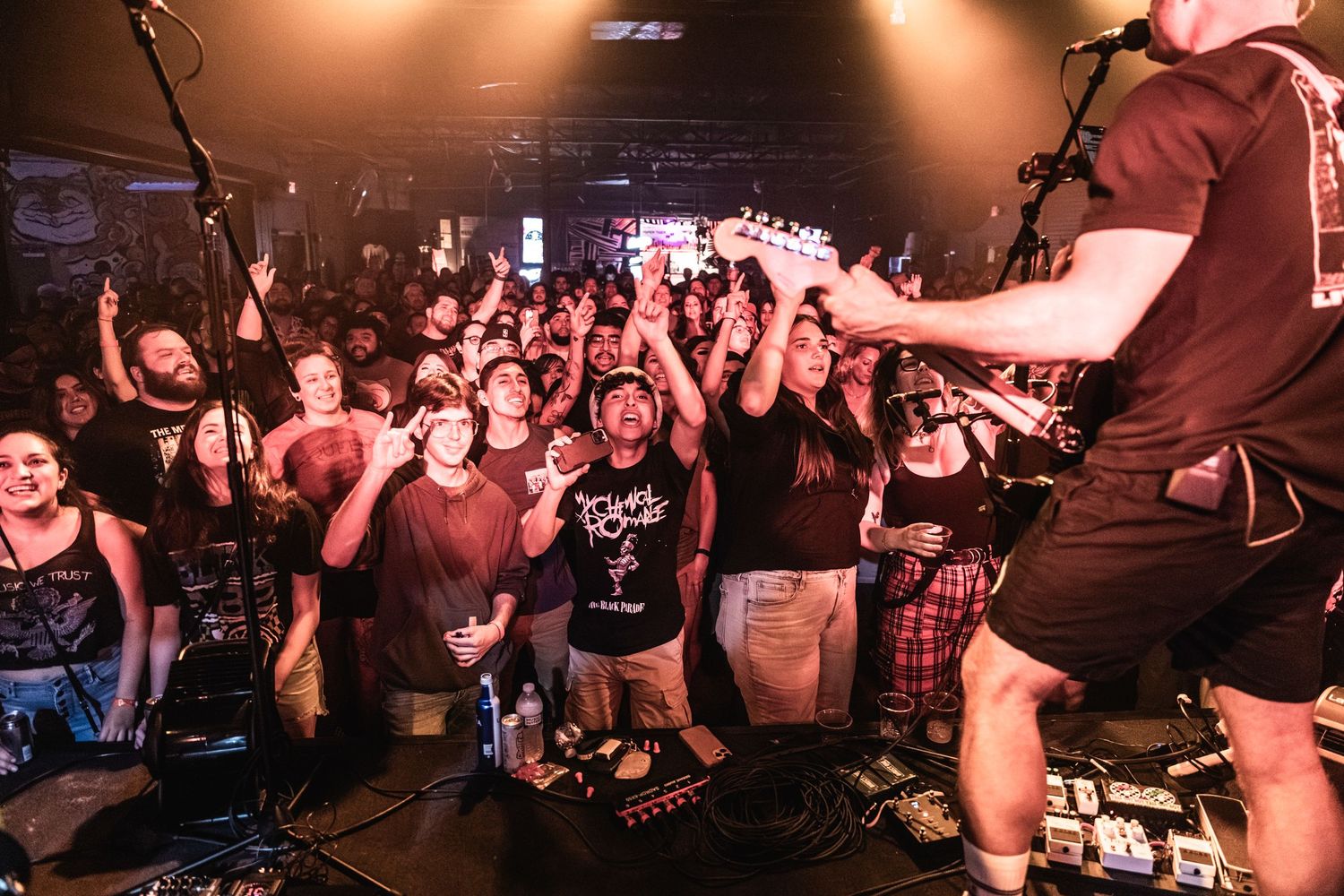 Energetic concert crowd cheers and raises their hands at a dimly lit music venue during a live performance.