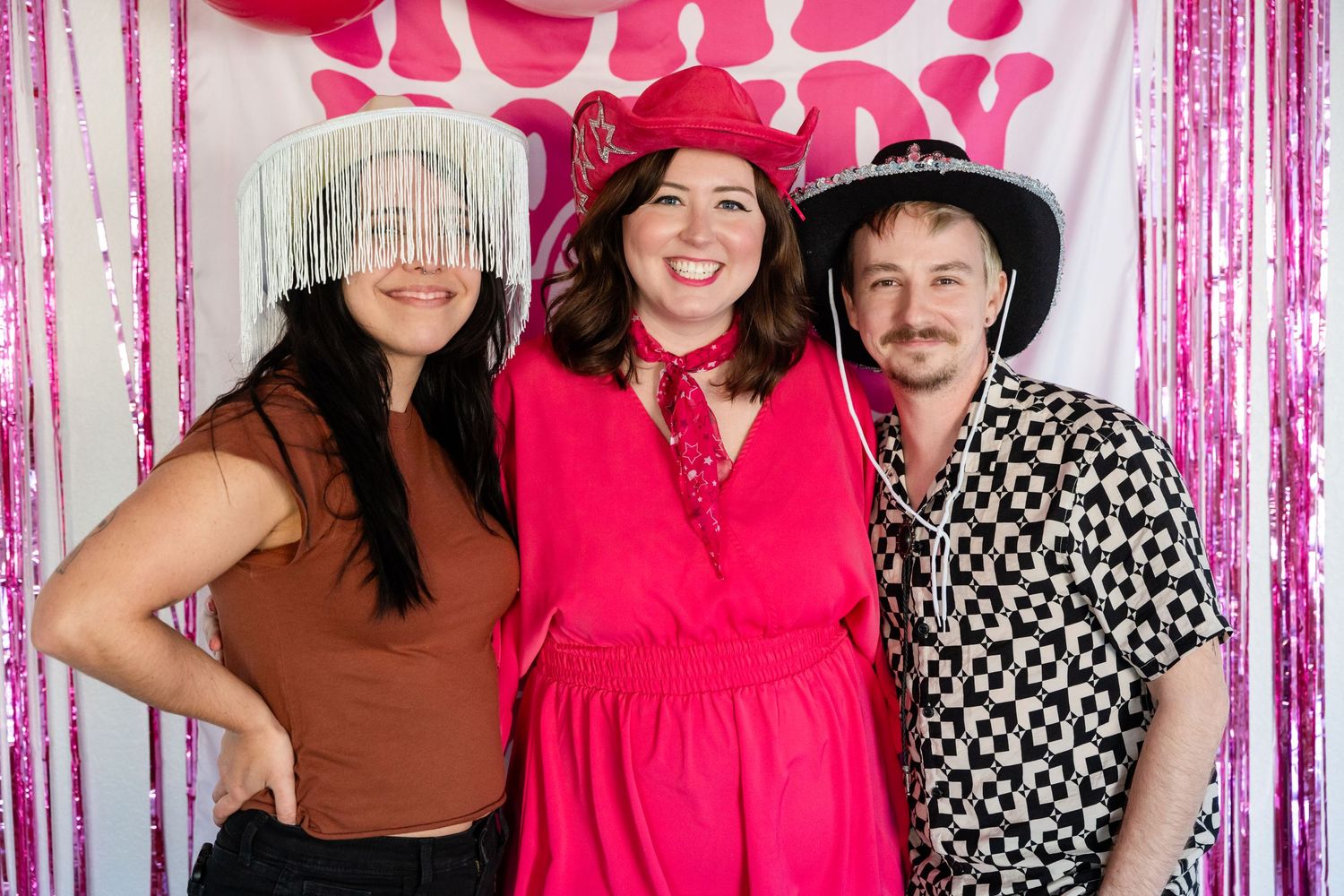 Three friends pose together at a photo booth with decorative fringe backdrop, wearing western style hats and festive attire.