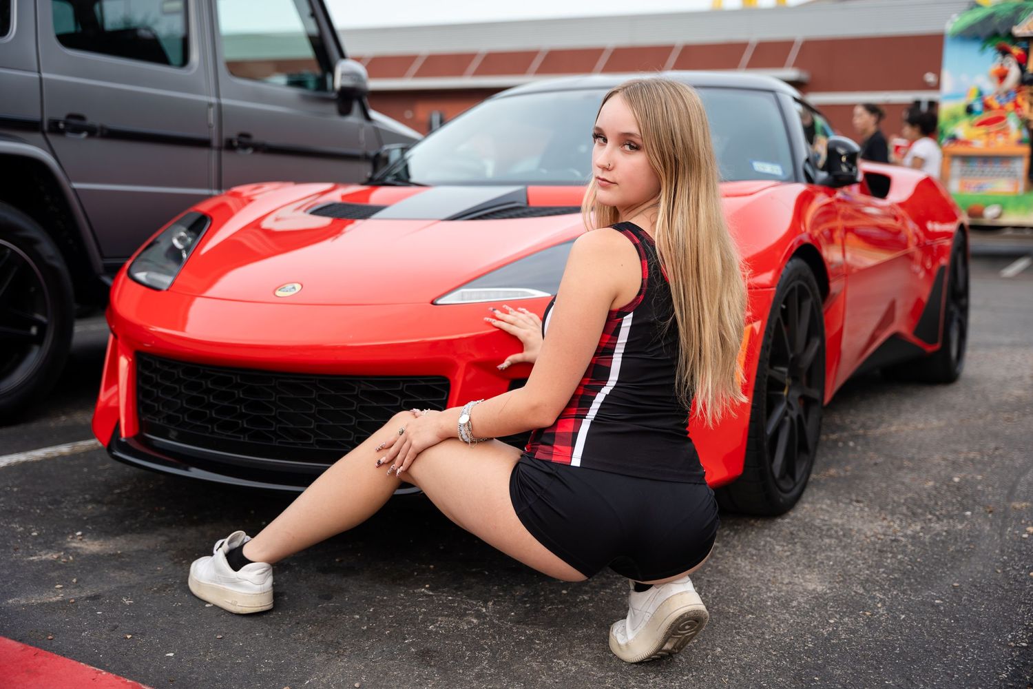 Model in black outfit sits posed next to bright red sports car in parking lot.