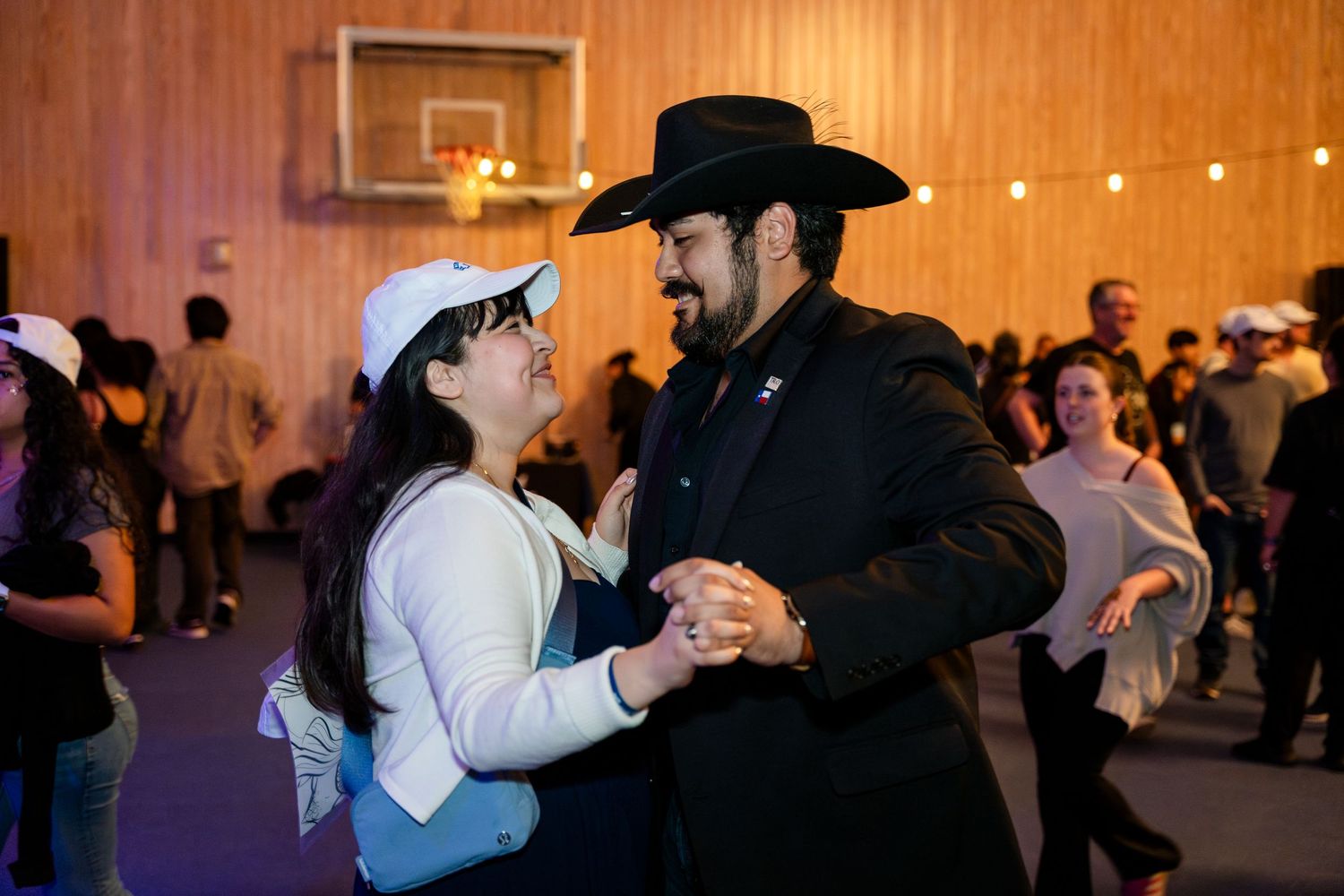 A couple dances together at an indoor event, with the man wearing a black cowboy hat and the woman in a white top.