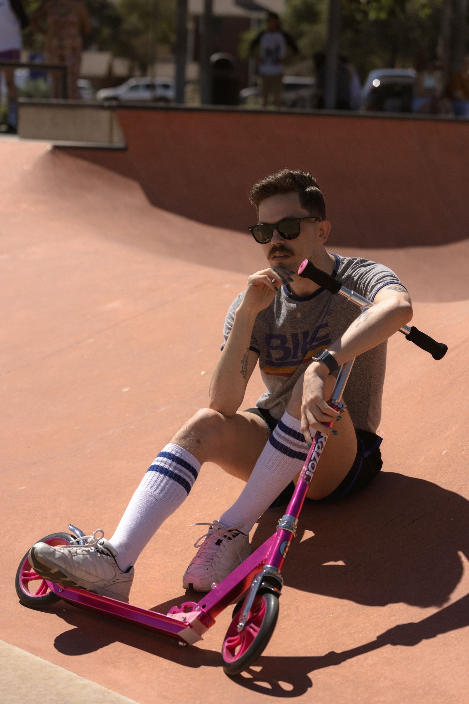 Person sitting with pink scooter at skate park wearing sunglasses and striped socks.