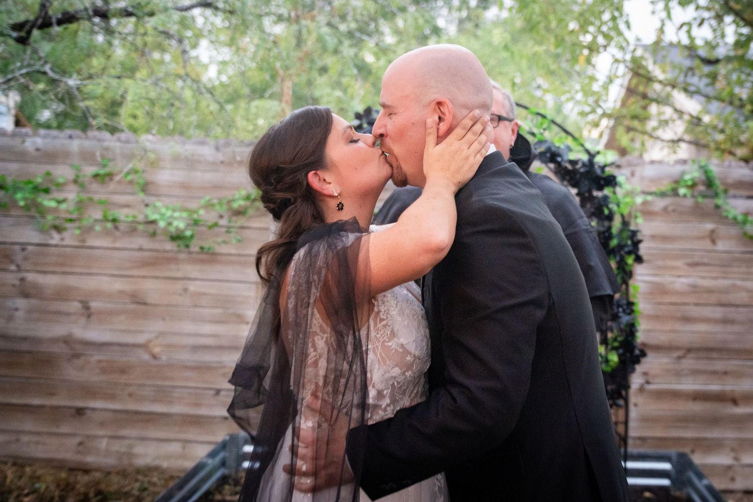 A romantic kiss between a couple at an outdoor wedding with wooden fence backdrop and greenery decorations.