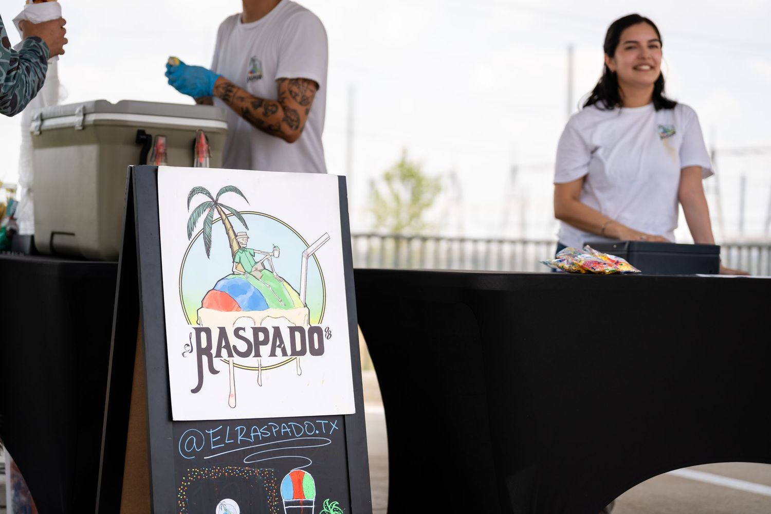 Food vendor booth with Raspado signage and staff preparing refreshments at an outdoor event.