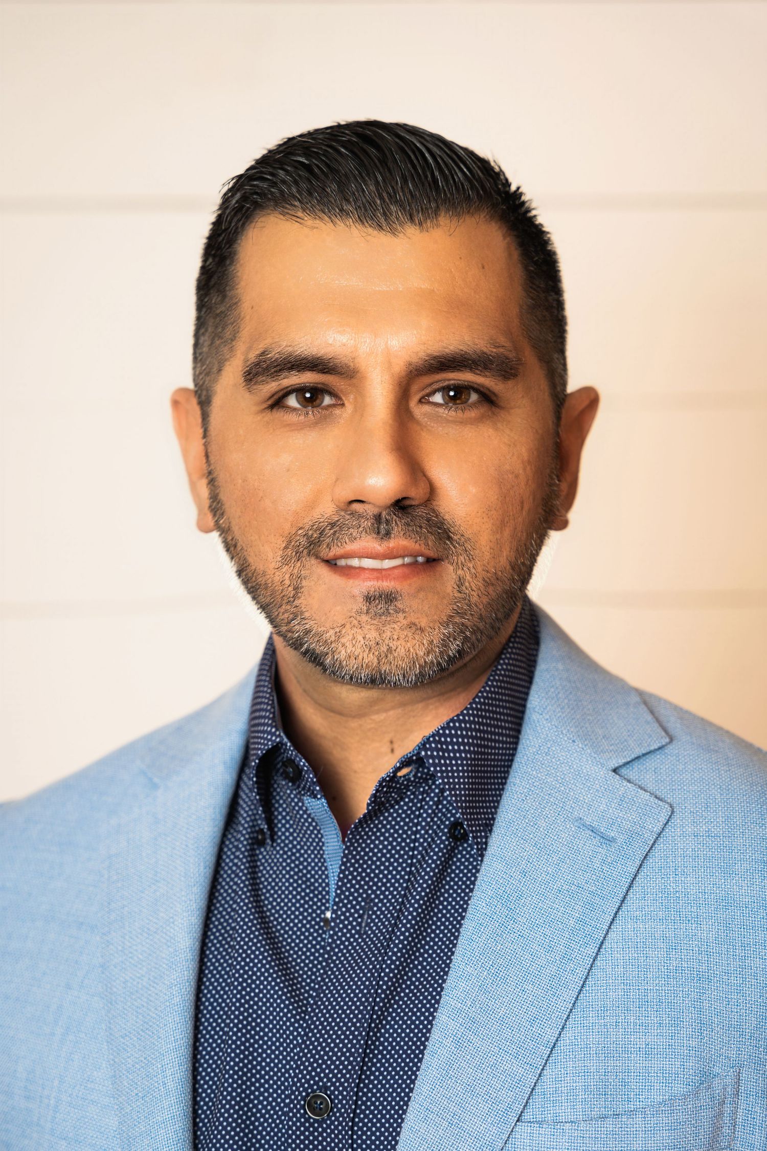 Professional headshot of a man in a light blue blazer and patterned shirt against a neutral background.