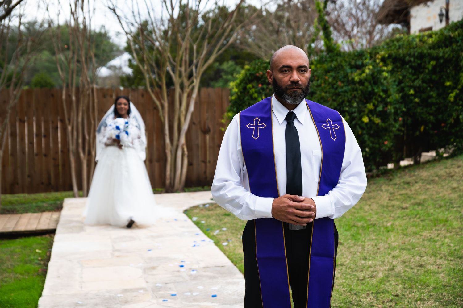 An officiant in purple robes stands on a pathway with a bride visible in the background.