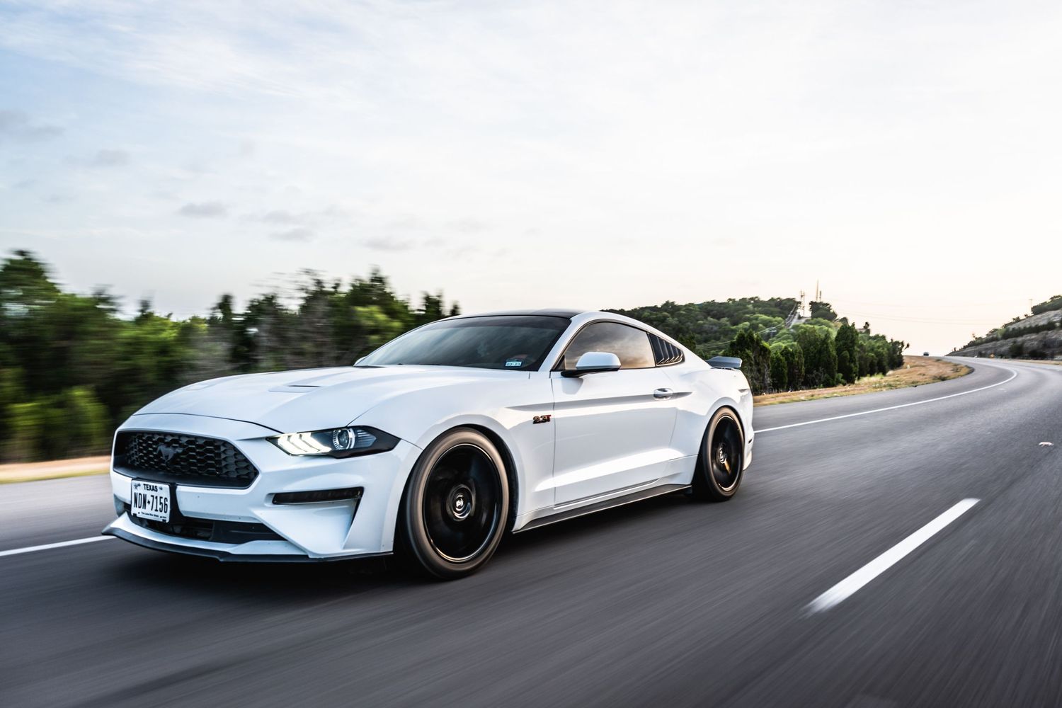 White Ford Mustang with black wheels photographed in motion on a highway.