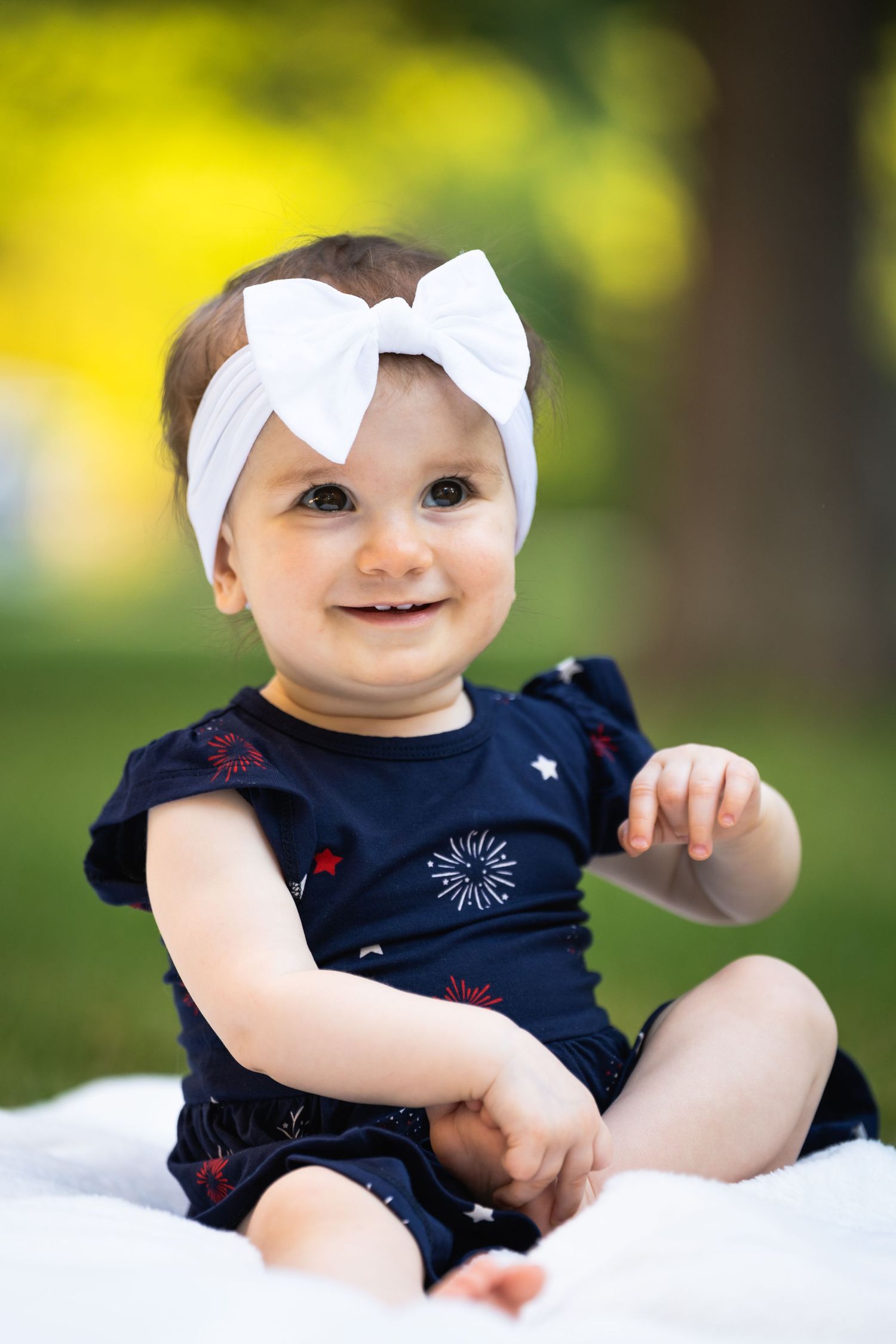 Baby wearing a navy floral outfit and white bow headband smiling outdoors on a sunny day.