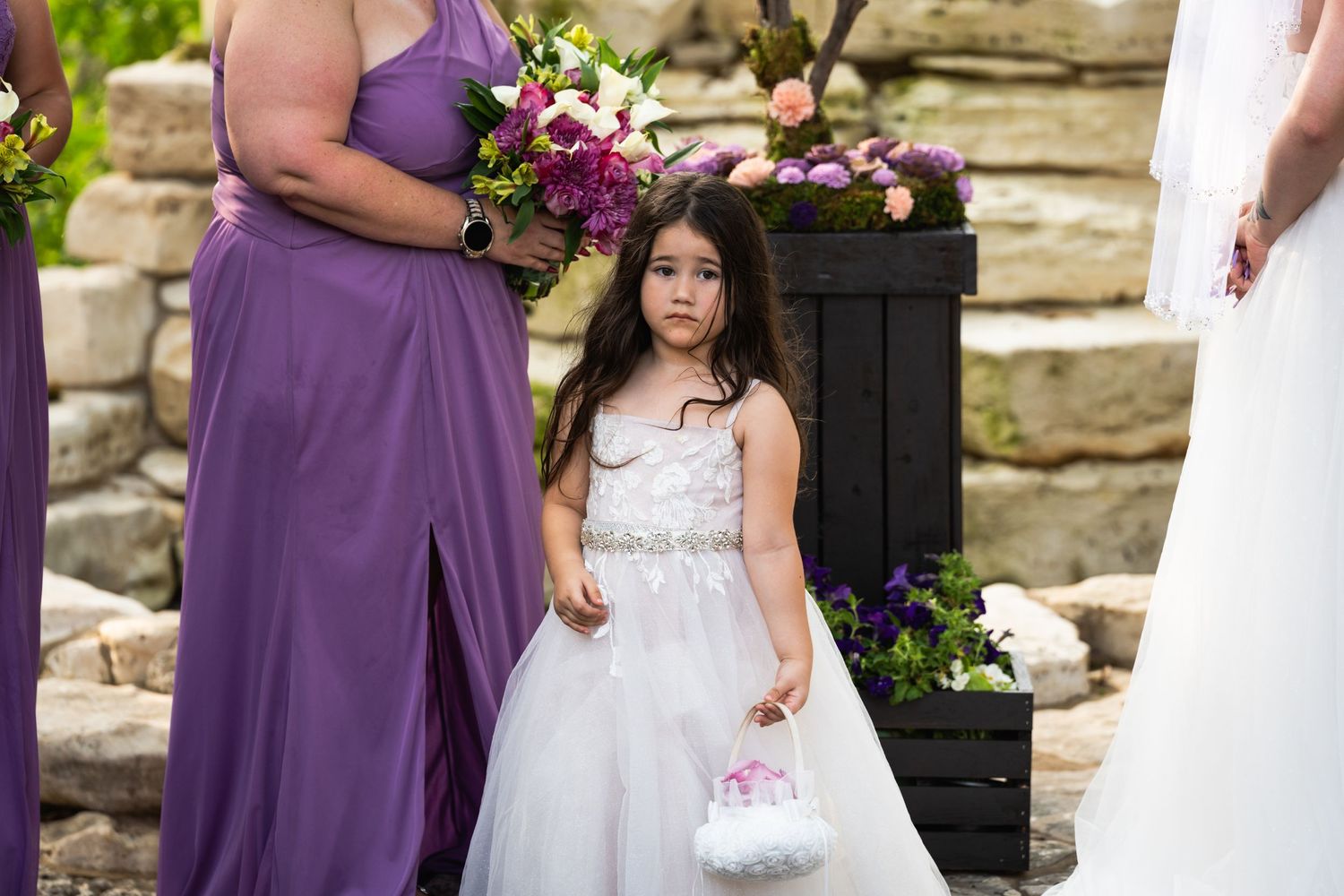 A flower girl in a white dress stands next to a bridesmaid wearing a purple gown at an outdoor wedding ceremony.