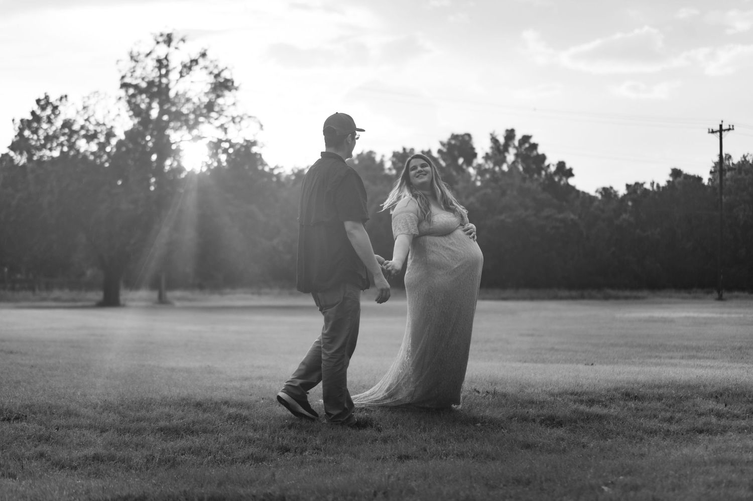 Black and white maternity photo of couple dancing in sunlit outdoor setting.