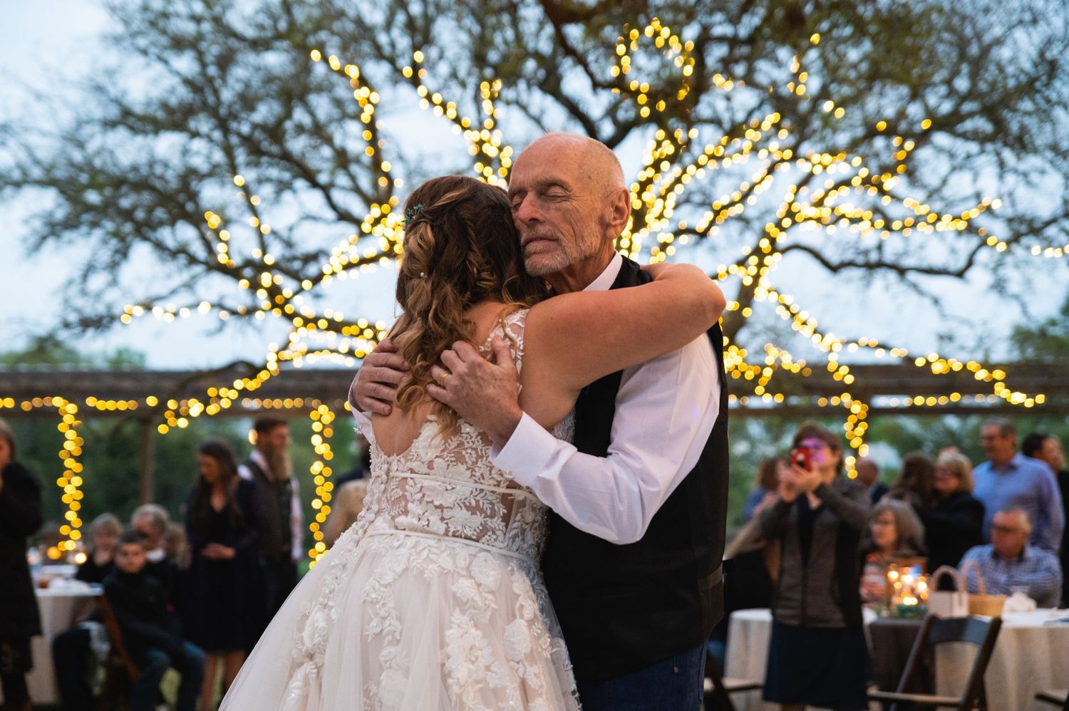 Couple embraces during a romantic dance with string lights and trees glowing in the background.