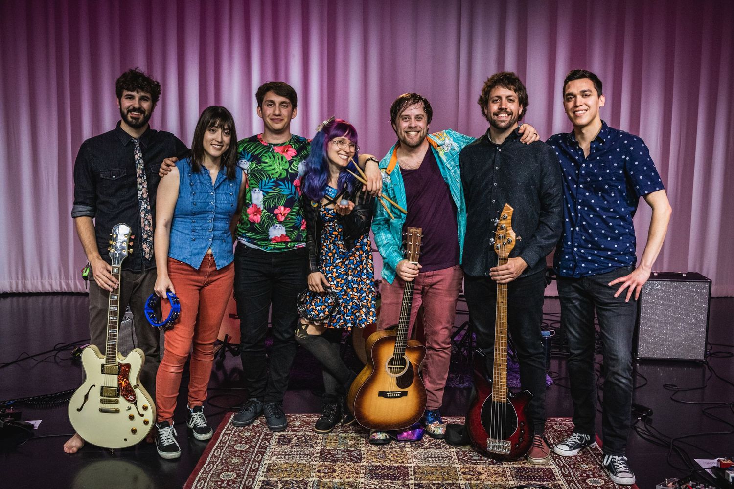 Musical group poses with their instruments on stage in front of pink curtain.