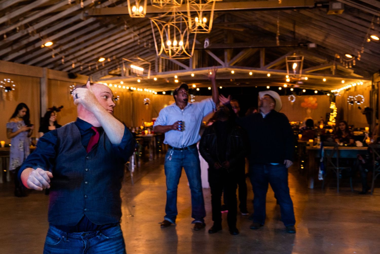 Wedding reception guests dancing under rustic chandeliers in a barn-style venue.