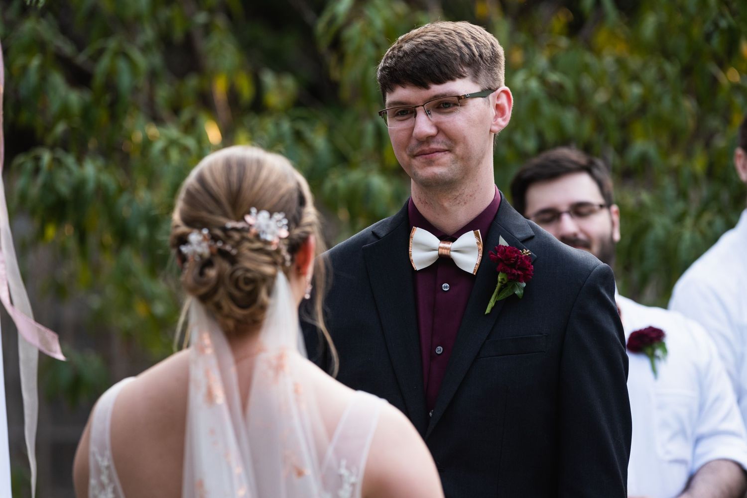 A wedding ceremony moment outdoors with attendants in formal attire against a leafy background.