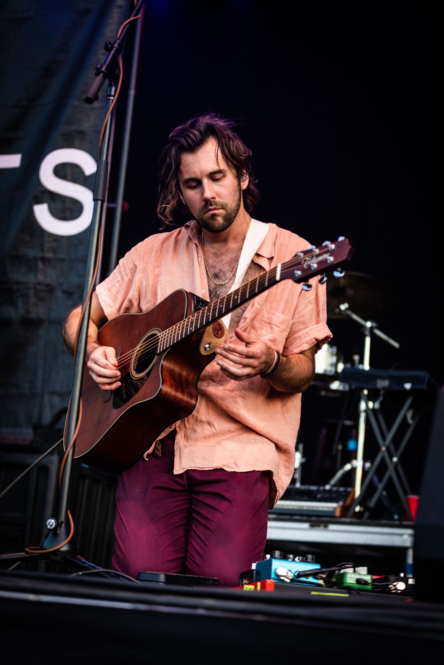 Concert performer plays acoustic guitar in pink shirt and burgundy pants on outdoor festival stage.