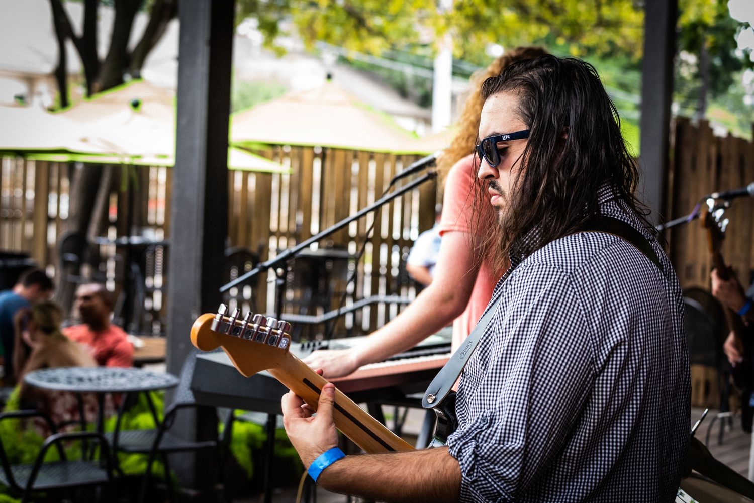 Musician plays guitar while singing at outdoor patio venue with tables visible in background.