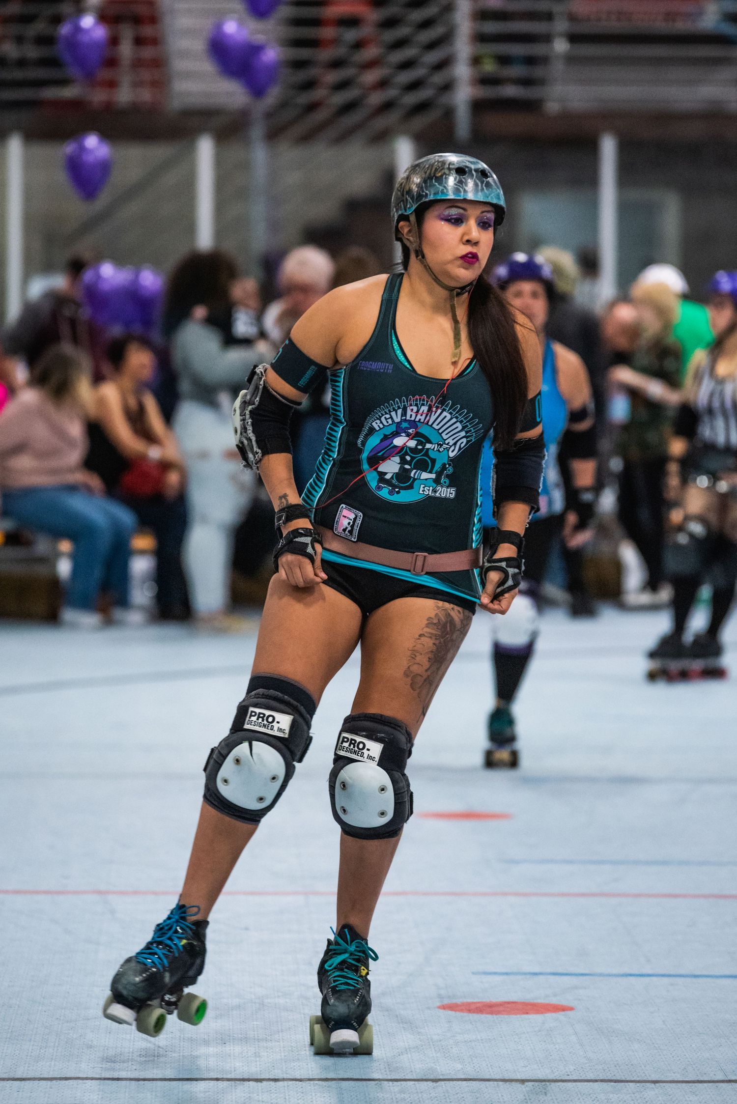 Roller derby athlete in blue uniform and protective gear skates on indoor rink with purple balloons.