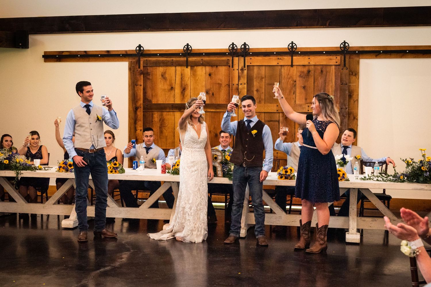 Wedding party members celebrate with raised hands during reception speeches at rustic venue.