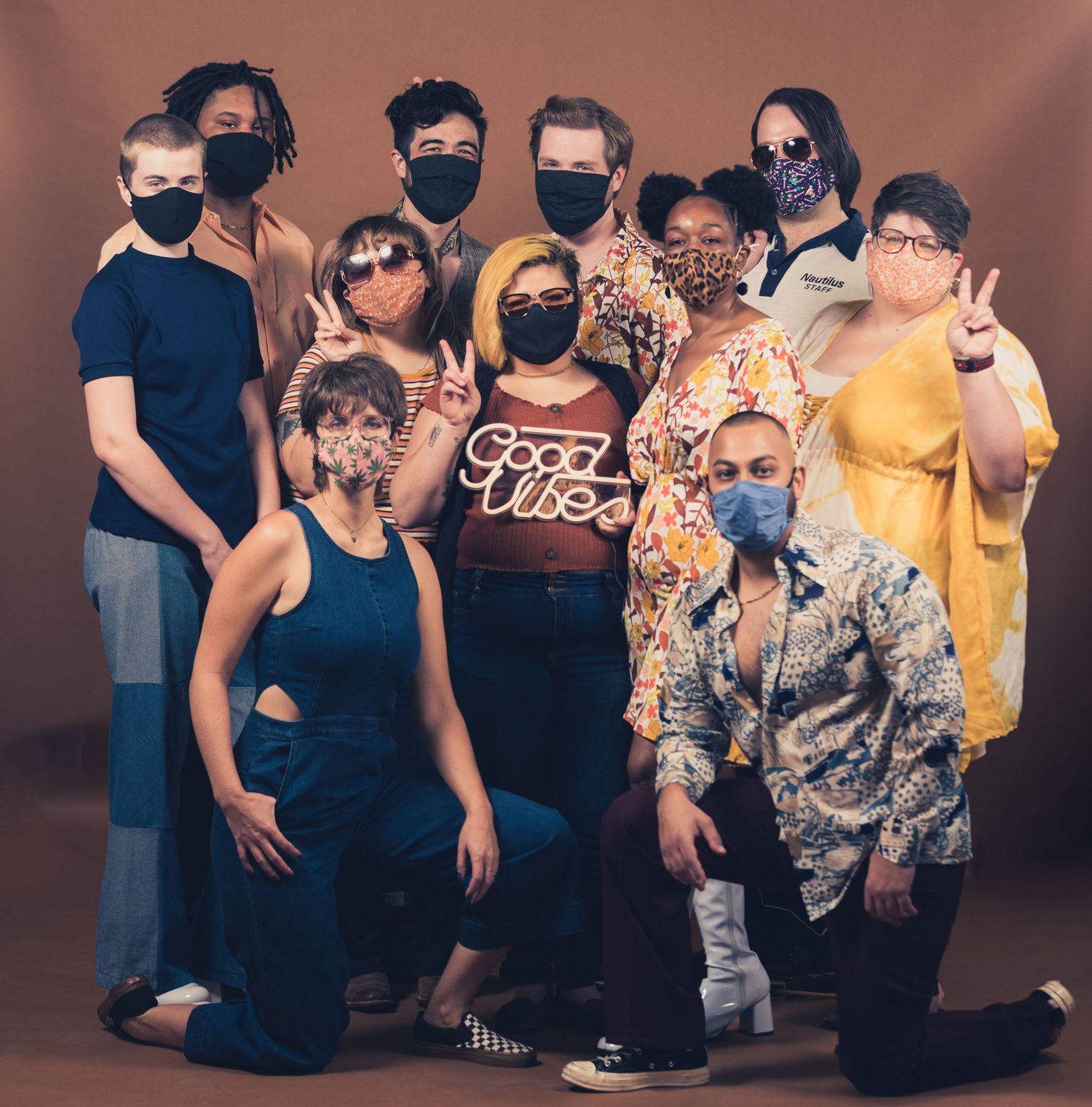 Group photo of people wearing masks posing together with peace signs and a Good Vibes neon sign.