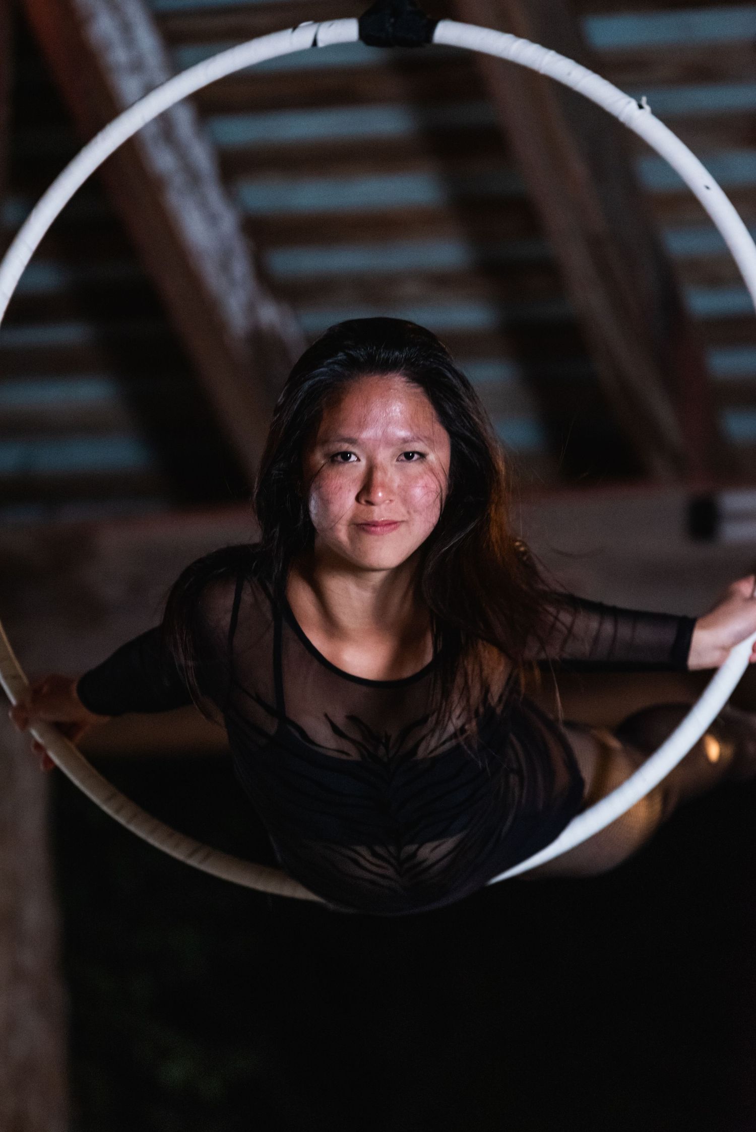 Aerial artist gracefully poses inside illuminated white hoop beneath wooden beams.