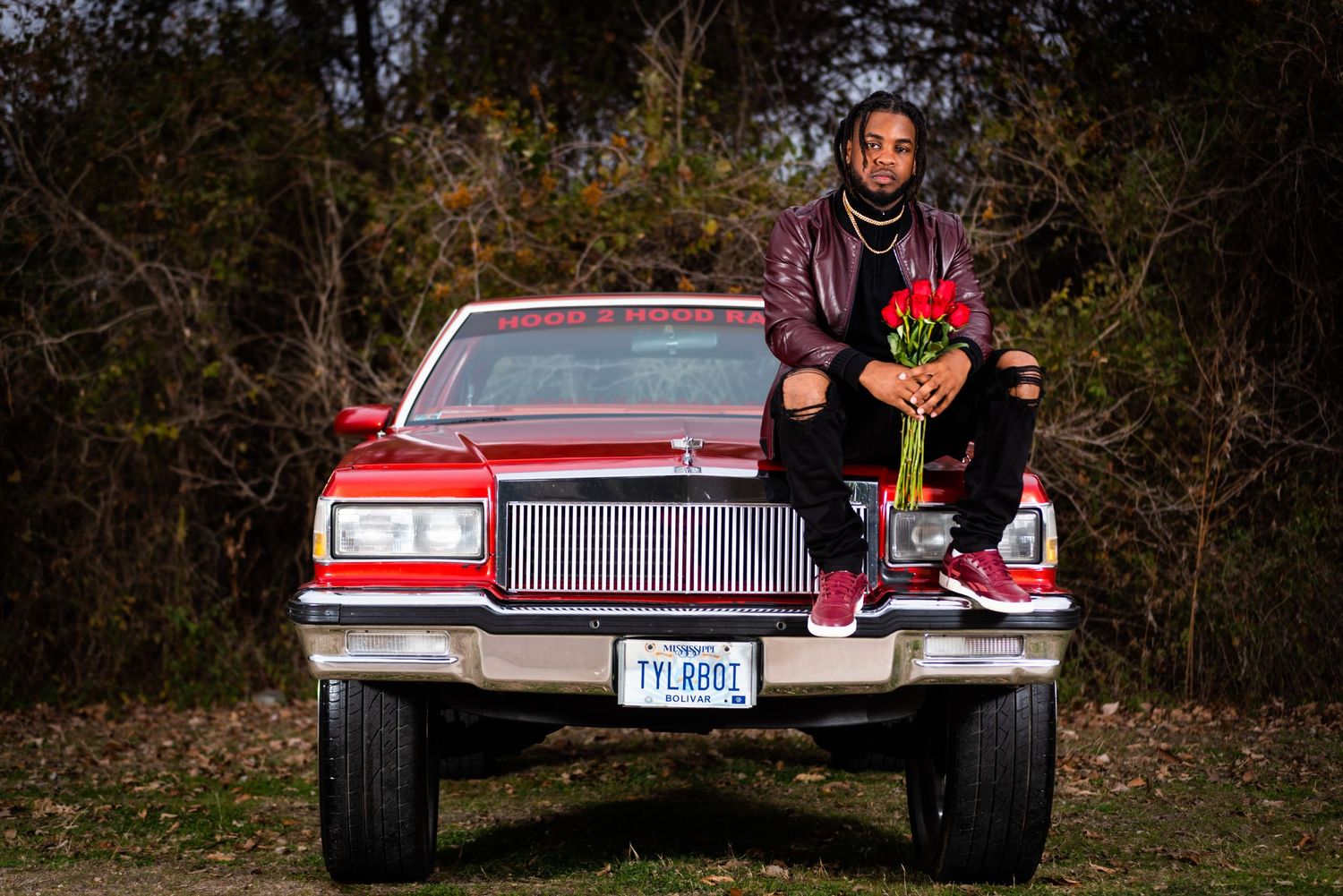 Person holding flowers while sitting on hood of vintage red car in natural setting.