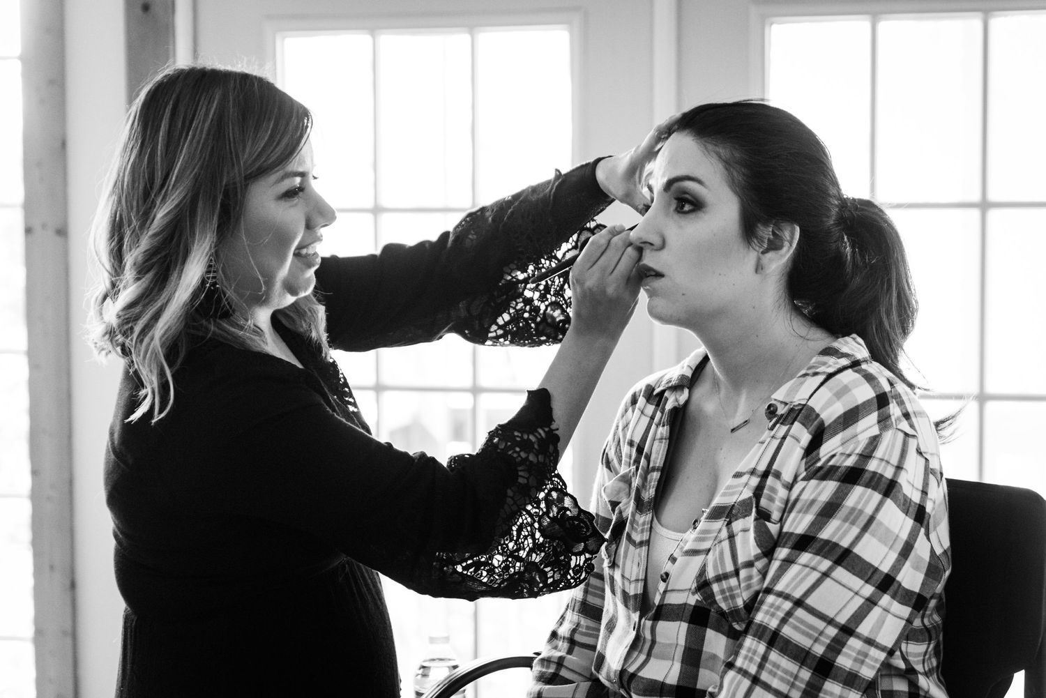 A makeup artist applies cosmetics in a bright room with windows.