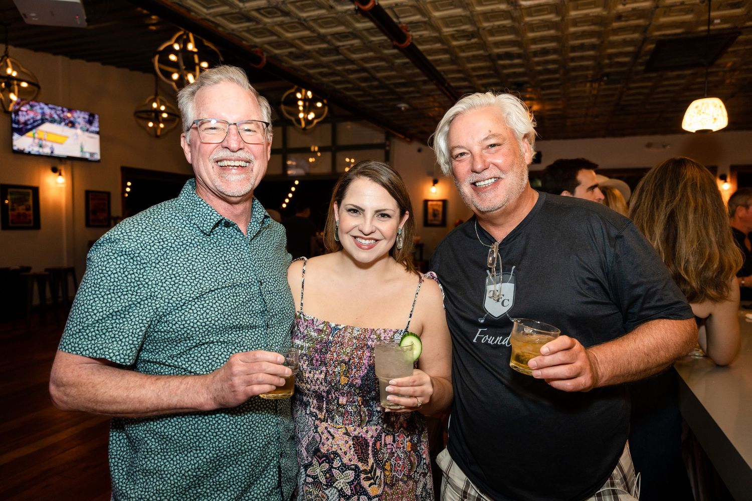 Three people smile while enjoying drinks together at an indoor venue with mood lighting.