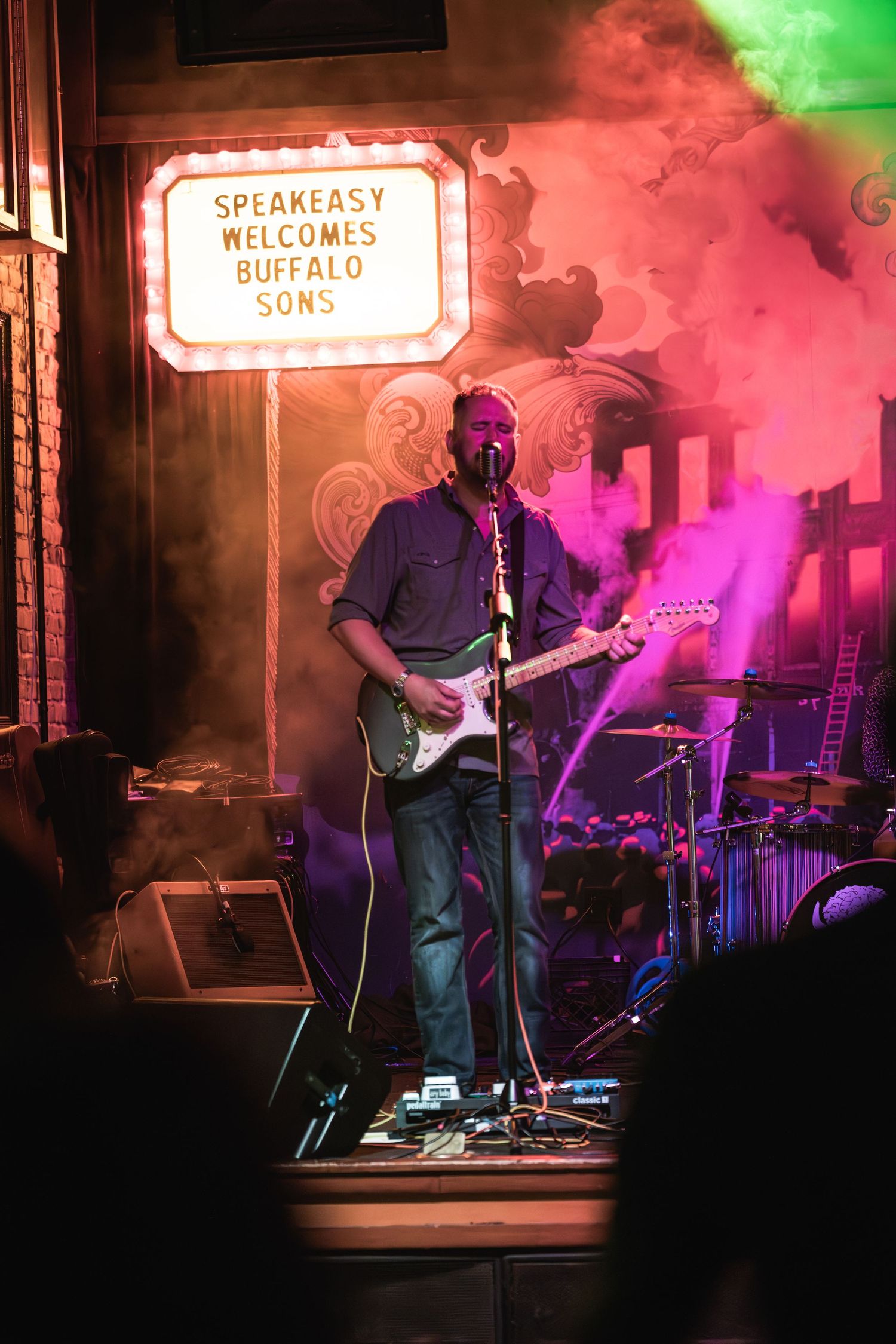 Musician plays guitar under colorful smoke effects with Speakeasy welcome sign visible above.