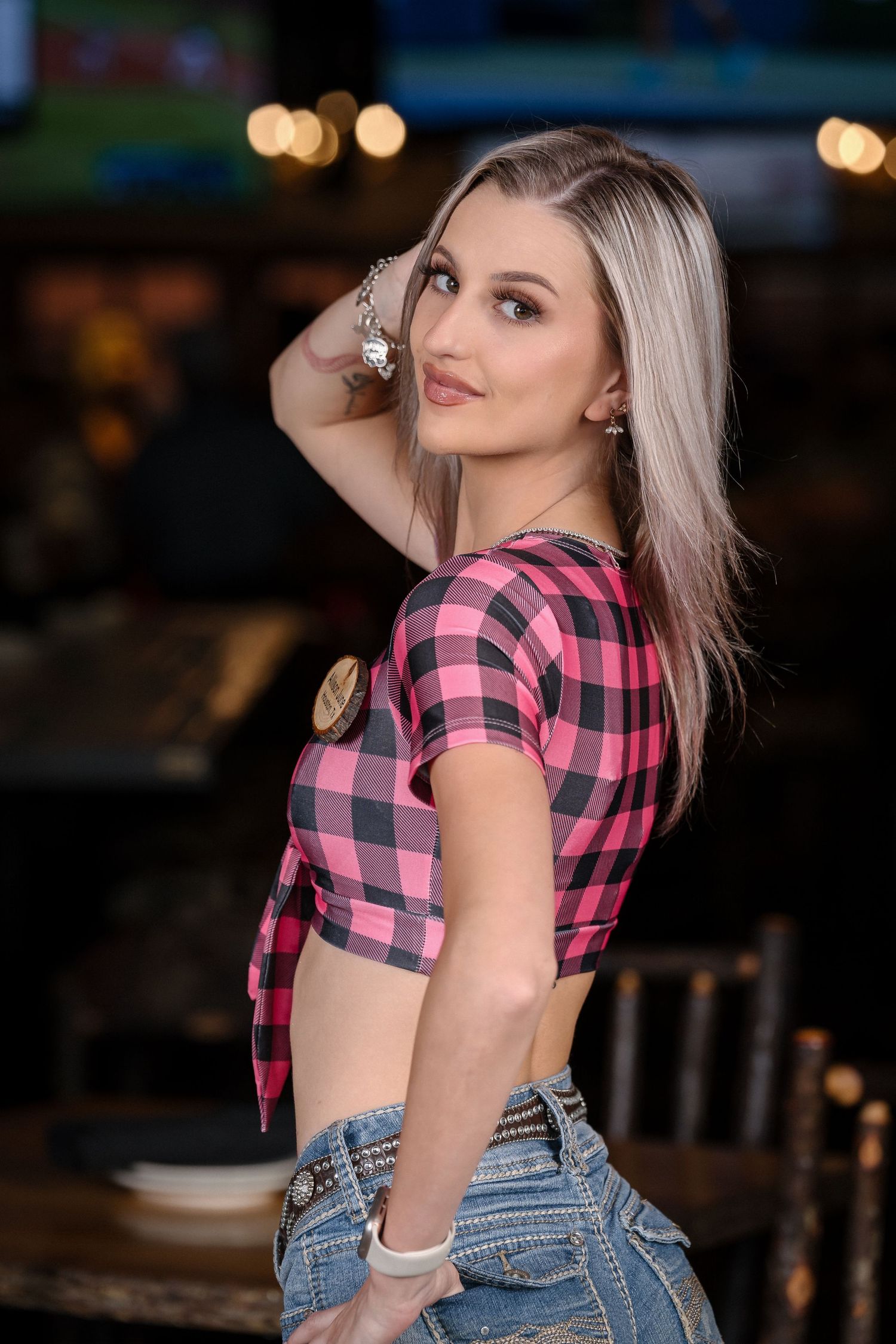 Model wearing pink plaid crop top and jeans poses against evening backdrop with bokeh lights.