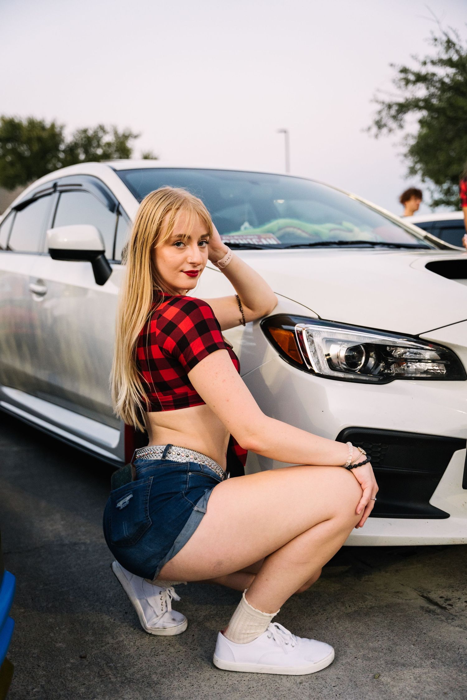 Person in red buffalo plaid crop top and denim shorts poses with silver Subaru WRX.
